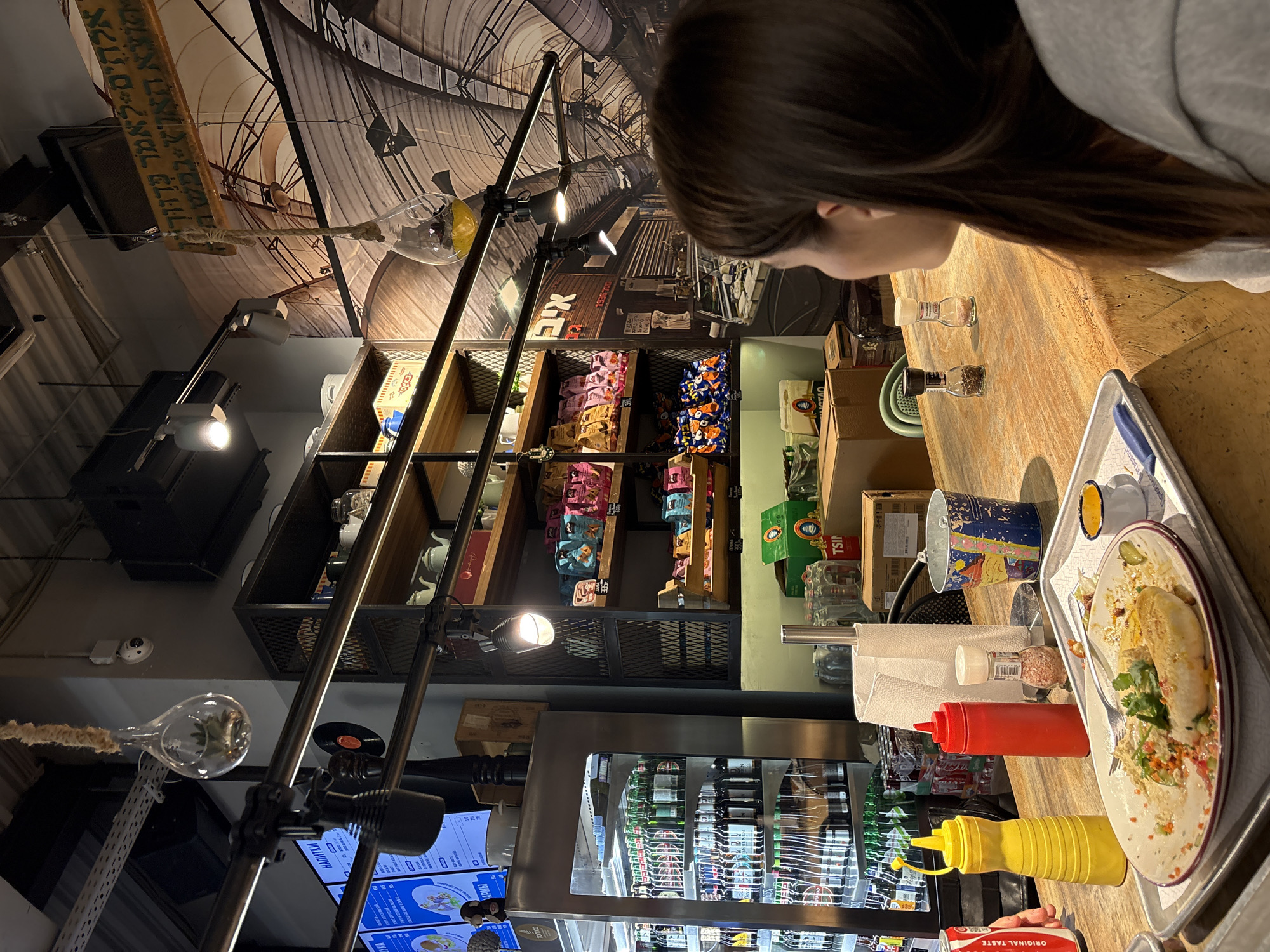 The image shows a woman with long, dark hair looking down at a plate of food on a wooden table. The plate contains a dish that appears to be a stuffed crepe or wrap, topped with fresh herbs and other ingredients. Next to the plate are several condiments, including squeeze bottles of yellow and red sauce, a salt shaker, and a pepper grinder.
Behind the woman and the table, there is a shelving unit filled with various items, including packaged snacks and boxes. A refrigerated display case with bottles of beverages is also visible.
The interior of the restaurant or cafe has an industrial-chic aesthetic, with exposed metal frameworks, pipes, and light fixtures. A large mural or wallpaper depicting a historical scene or cityscape covers the wall above the shelving unit. There are also some Hebrew inscriptions on a wooden board, indicating the location may be in Israel.
Based on the details available, it is not possible to determine the location in the city of Moscow, Russia. The details suggest the photo was taken in Israel.