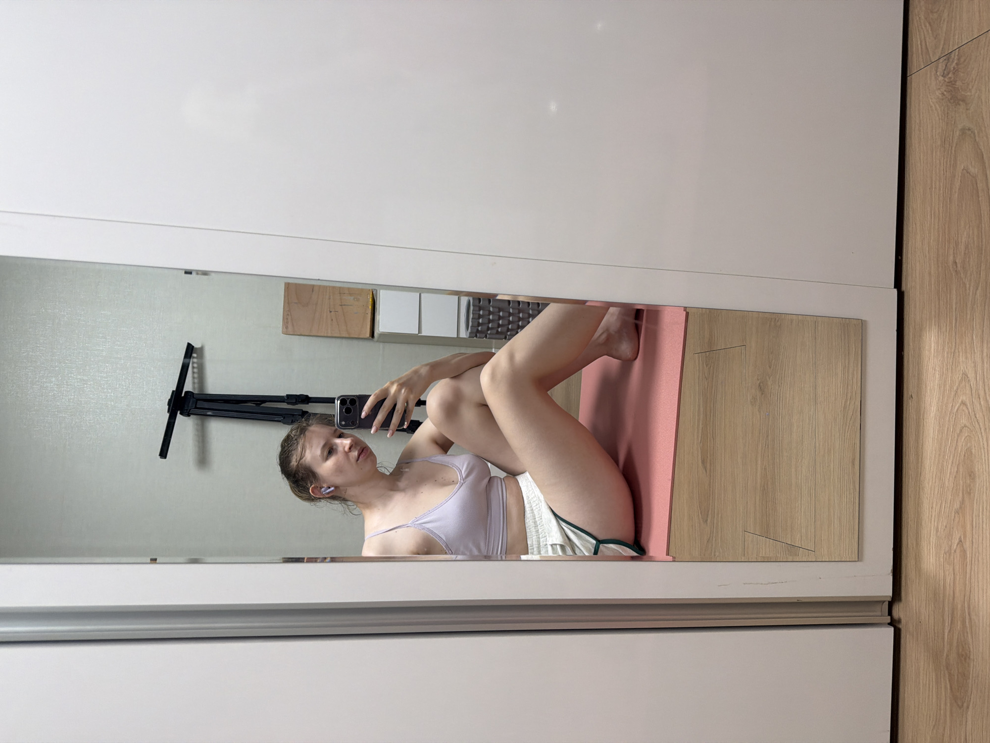 A young woman with blonde hair pulled back in a bun, wearing a light purple sports bra and light-colored athletic shorts with green trim, sits on a pink yoga mat. She is positioned on the floor, facing a mirror and holding a smartphone to take a selfie. The woman is wearing light purple wireless earbuds. The background of the mirror reflection shows a wall with a textured wallpaper, a black tripod with a mounted light, and some wooden and white shelves. The room appears to be a bedroom or living space with wooden flooring visible. The woman is seated in a relaxed pose, with her legs bent and crossed. The overall scene suggests a casual, indoor setting, possibly a home gym or a personal workout space.