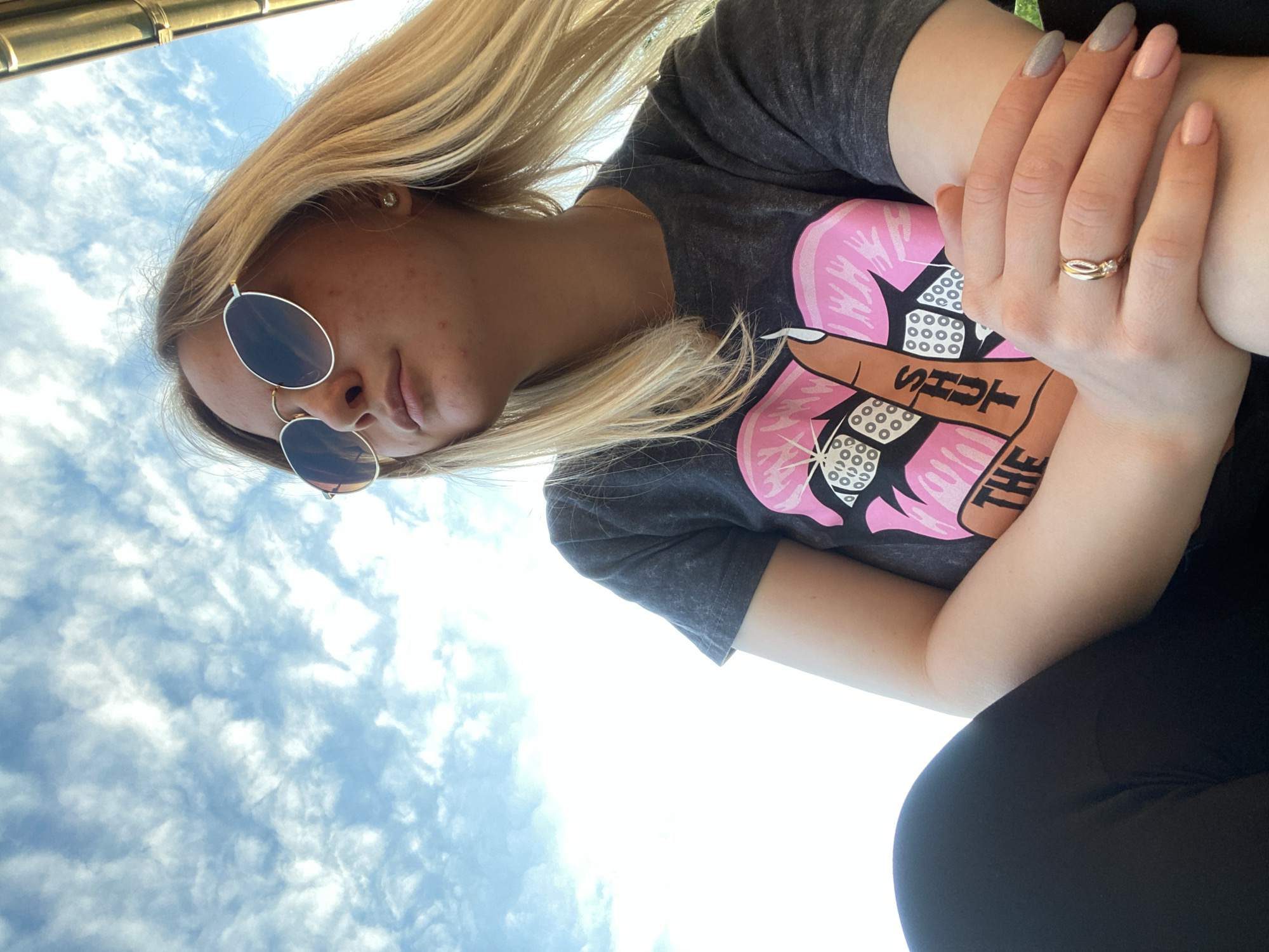 A young adult female with long blonde hair and sunglasses is depicted in a low-angle outdoor shot against a bright blue sky with scattered white clouds, in the city of Unknown, Unknown. She wears round, gold-rimmed sunglasses with dark lenses. Her facial skin shows minor blemishes, and a small stud earring is visible in her right ear. She is wearing a dark grey t-shirt with a pink and brown graphic print of a hand making a middle finger gesture, with the words "SHUT THE" visible on the fingers. Her arms are crossed, with her left hand resting over her right forearm. Her fingernails are manicured with both light pink and grey polish. On the ring finger of her left hand, she wears two gold-toned rings: one with a clear gemstone in a twisted band design, and a simpler inner band. Her lower body is clad in black leggings or pants, visible in the lower left. In the upper right corner, a curved metallic structure, possibly part of a building or awning, is partially visible. The lighting is bright, indicating daytime.