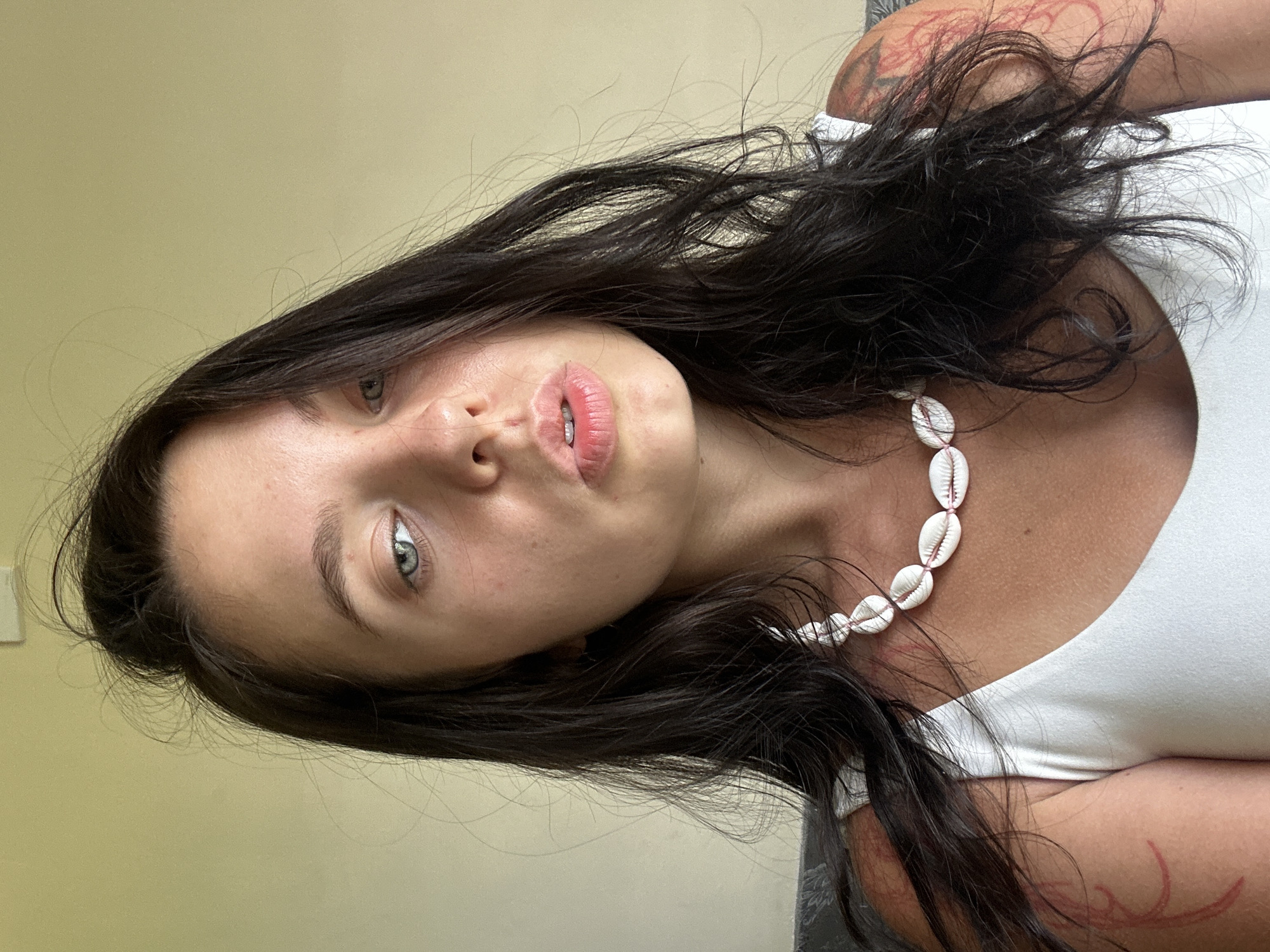 A young woman with dark, wavy hair and light blue eyes poses for a close-up selfie against a light yellow, textured wall. She is wearing a white tank top and a necklace made of small white seashells strung together with pink cord. Her left arm is visible, and it appears to have a red tattoo or temporary marking. The woman's mouth is slightly parted, and her gaze is directed towards the camera. The lighting is bright, suggesting an outdoor or well-lit indoor environment.