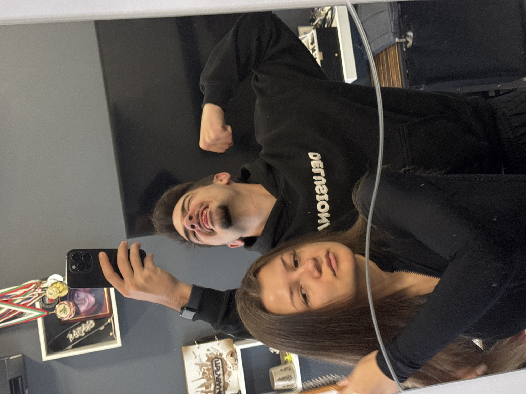 The image shows a selfie taken in a mirror. A young man with a beard is flexing his arm, wearing a black hoodie with white lettering. A young woman with long brown hair is standing beside him, holding a hairbrush. Both are looking at the camera, which is held by the man. The background includes a grey wall, a black panel, shelves with objects, and what appears to be a framed picture with medals and a skateboard. There is also a thin white cord reflecting in the mirror. The location cannot be determined definitively as Sofia, Bulgaria, from the available details.