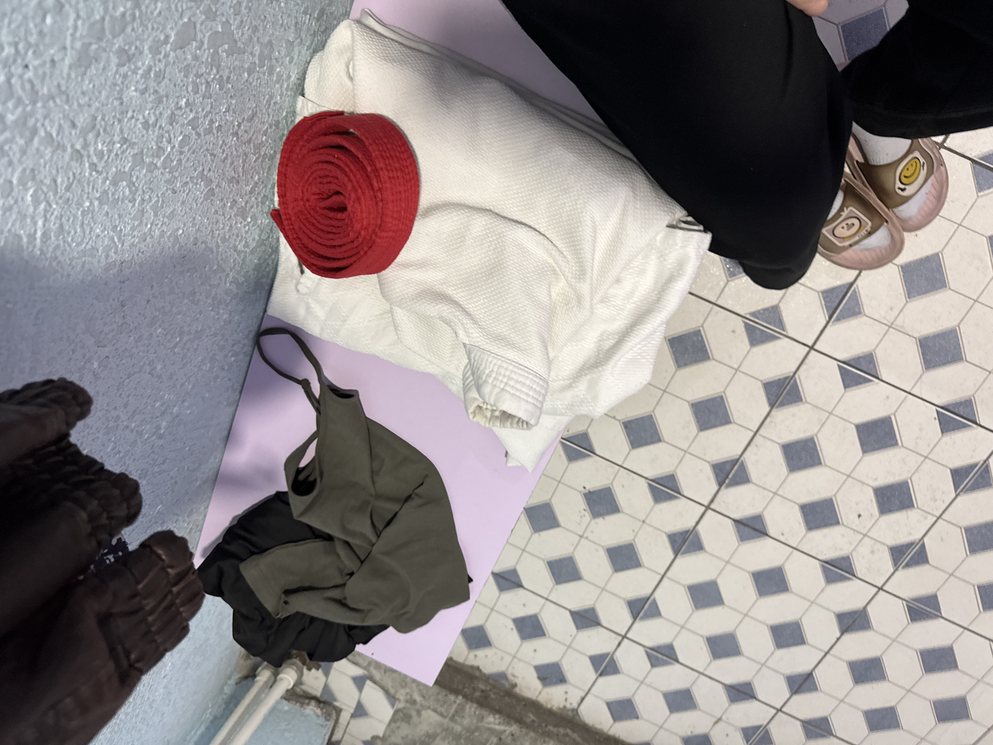The image shows a top-down view of various objects on a lavender surface and a tiled floor. A rolled red martial arts belt sits next to a folded white martial arts uniform (gi). A pile of clothing, including an olive green tank top and black garments, is also on the lavender surface. In the upper right, a person wearing black pants and smiley-faced shoes stands on the tiled floor, which has a geometric pattern of white tiles with blue accents. In the bottom left corner, part of a brown puffer jacket is visible. The room is indoors, with textured light blue walls in the background. There is no verifiable information to suggest the image was taken in Yerevan, Armenia.