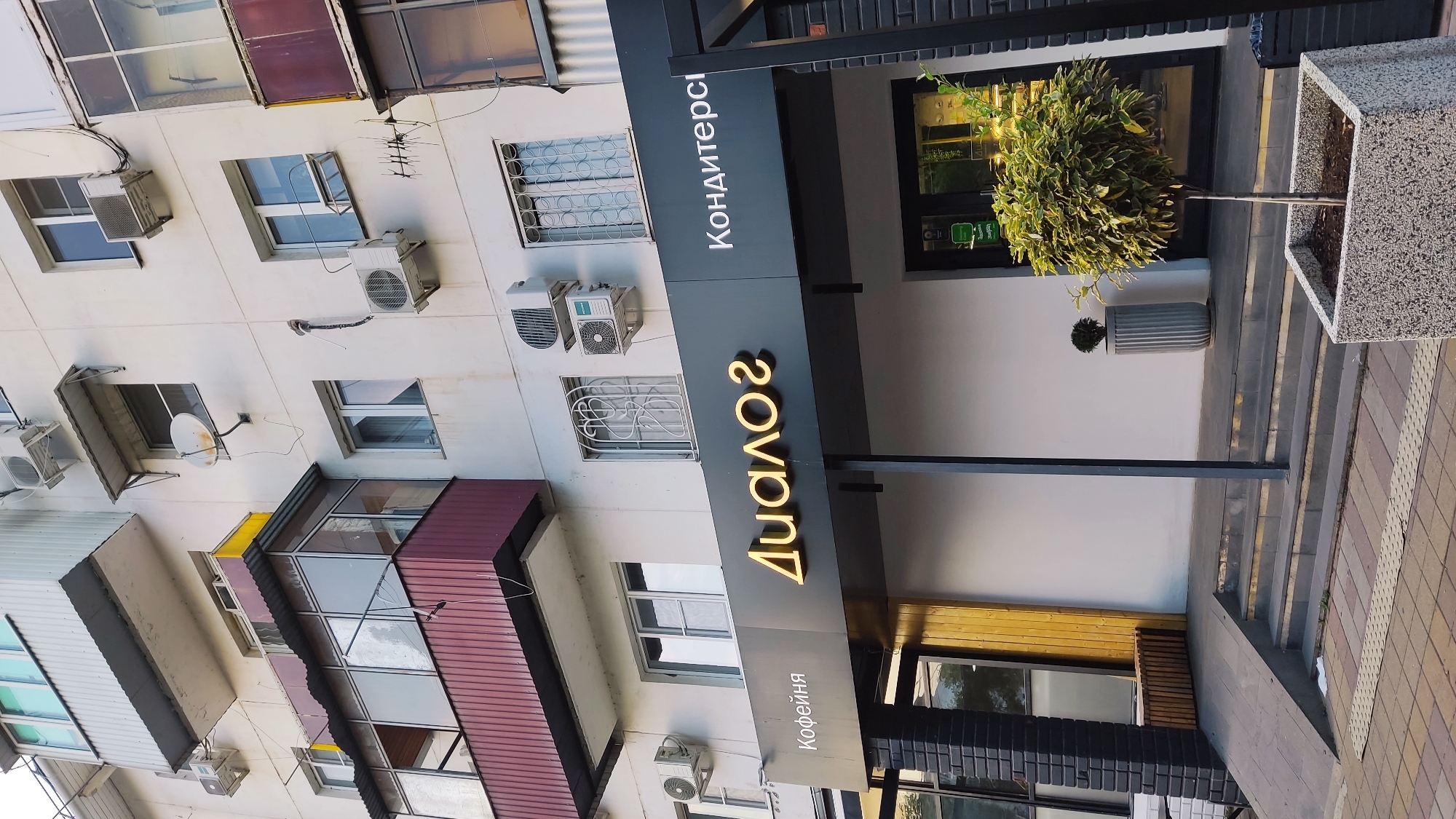 The image shows the entrance to a cafe and confectionery called "Dialog" in Krasnodar, Russia. The building is a multi-story apartment building with several balconies and air conditioning units visible. The cafe has a dark gray awning with illuminated text in Russian, reading "Coffee shop" on the left and "Confectionery" on the right. The central part of the awning features the illuminated name "Dialog" in large golden letters. Below the awning, the entrance is marked by a black column and a wooden facade. A ramp and stairs lead up to the entrance. There are planters with trees and a decorative pot outside the cafe. The overall impression is that of a modern establishment situated within an older residential building.
