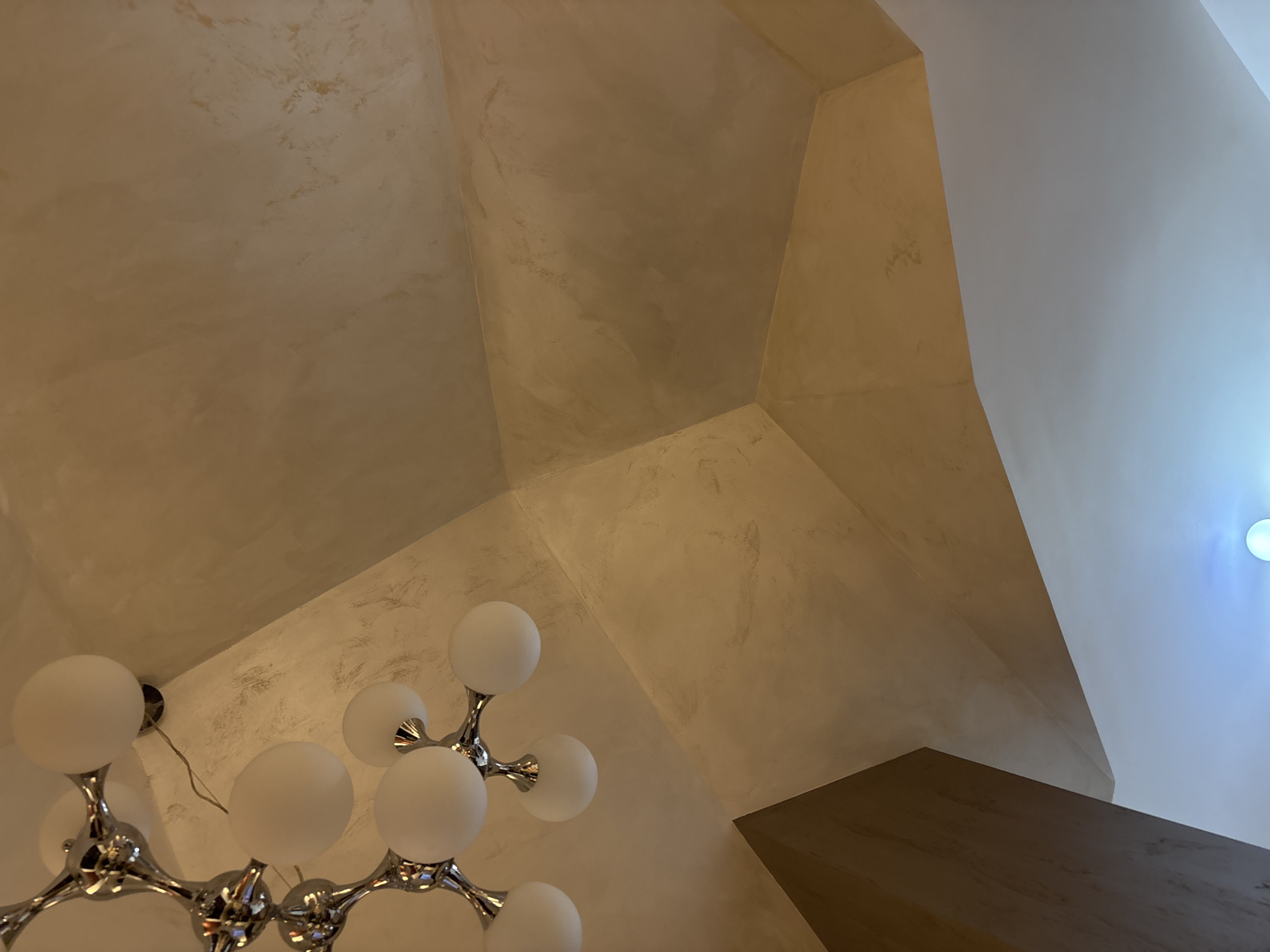 A low-angle shot of an interior ceiling with a modern chandelier. The ceiling has a multi-faceted design with varying shades of tan and white. The chandelier is chrome with spherical white bulbs. A portion of a dark wooden structure is visible in the lower right corner. The specific location in Plovdiv, Bulgaria, cannot be determined from the image.