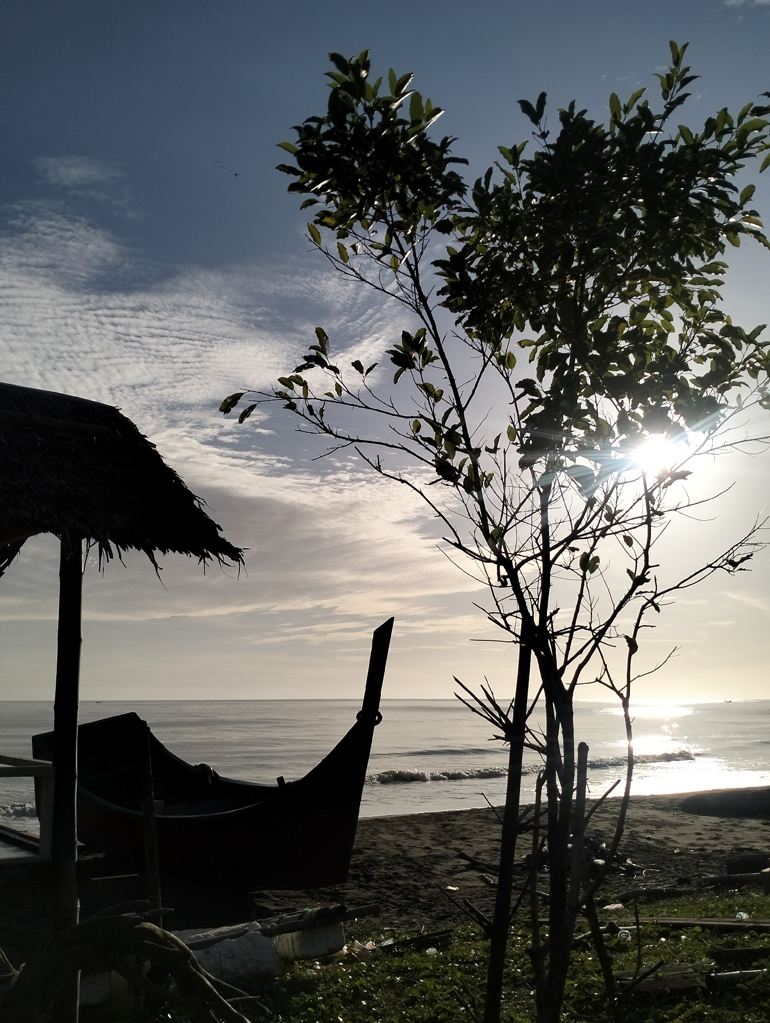 The image captures a beach scene in Sagoe, Indonesia. In the foreground, there's a silhouetted wooden boat nestled under a thatched roof structure. To the right of the boat, a tree with sparse leaves is silhouetted against the bright sunlight reflecting off the ocean. The beach stretches towards the calm water, and the sky is partly cloudy. There are no people visible in the scene. The image has a stark contrast, with the foreground appearing dark and the background, especially the ocean, brightly lit.