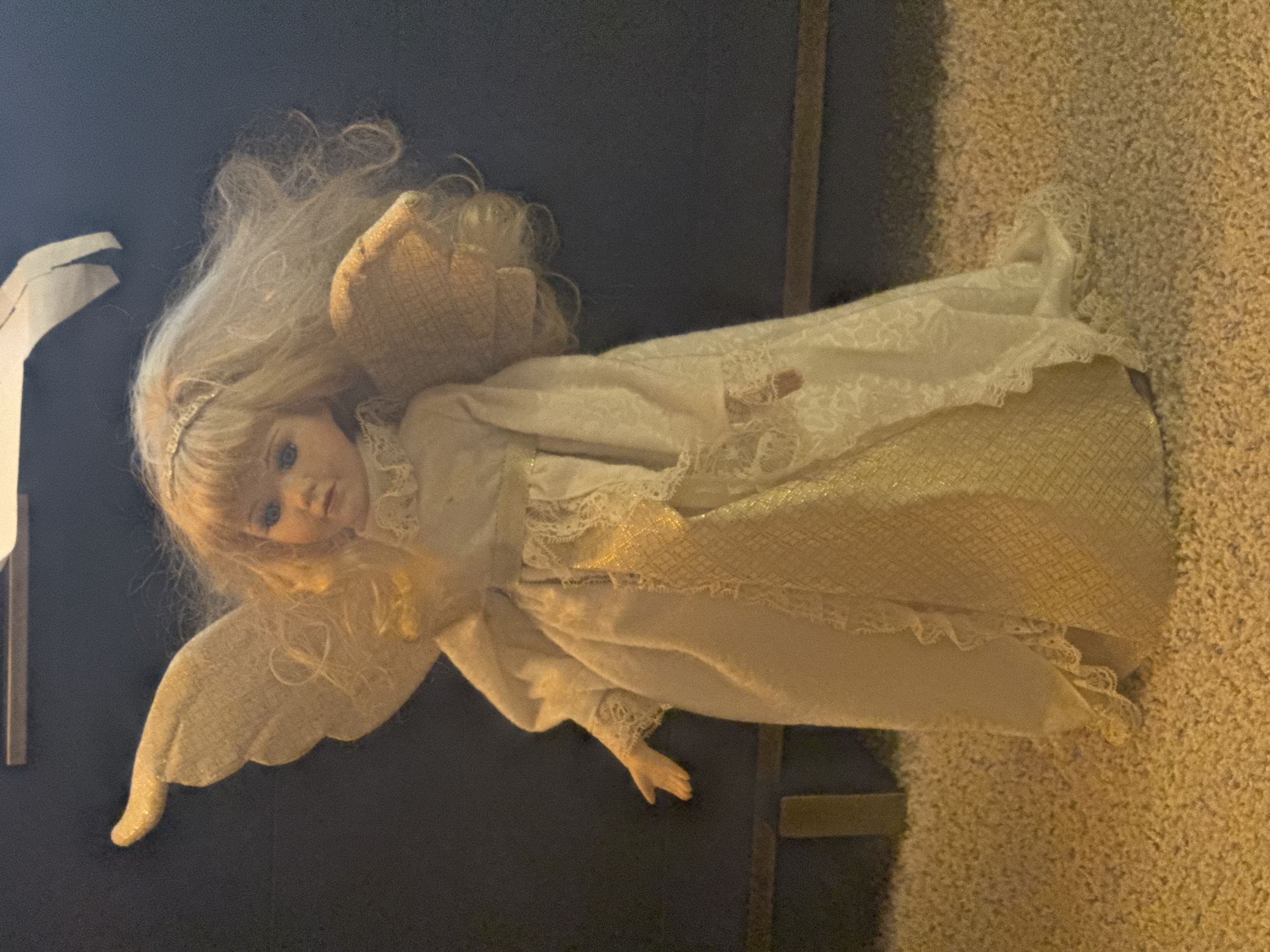 A porcelain doll, dressed as an angel, stands upright on a textured carpet. The doll has blonde hair styled with gentle waves, and a silver-toned headband adorns its head. Its eyes are blue, and its lips are painted a soft pink.
The angel costume consists of a flowing, off-white gown with lace trim and a gold-patterned insert down the front. The gown features long sleeves with lace cuffs. Attached to the doll's back are two large, textured wings, one on each side, and another smaller wing or accent is positioned on its right side. The doll's left arm is slightly bent outwards, with its hand open.
The background is a dark, solid color, possibly a wall or fabric, with a horizontal wooden element visible behind the doll. The lighting appears to be artificial, casting a warm glow on the doll and the carpet. The overall setting suggests an indoor environment.
There are no people present in the image, and no actions or interactions are depicted beyond the static pose of the doll. The image is devoid of any contextual clues that would place it within the city of Citrus Heights, United States of America.