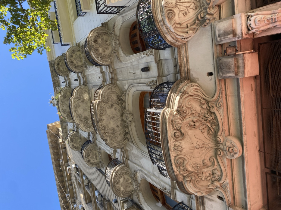 An upward angle captures the façade of an ornate building, adorned with protruding balconies. Each balcony is enclosed by black metalwork and topped with elaborate carvings. Sunlight illuminates the cream-colored stonework, showcasing intricate details in the sculptural relief. The sky is visible on the left. A green leafy tree appears in the upper left corner, adding a touch of natural contrast.