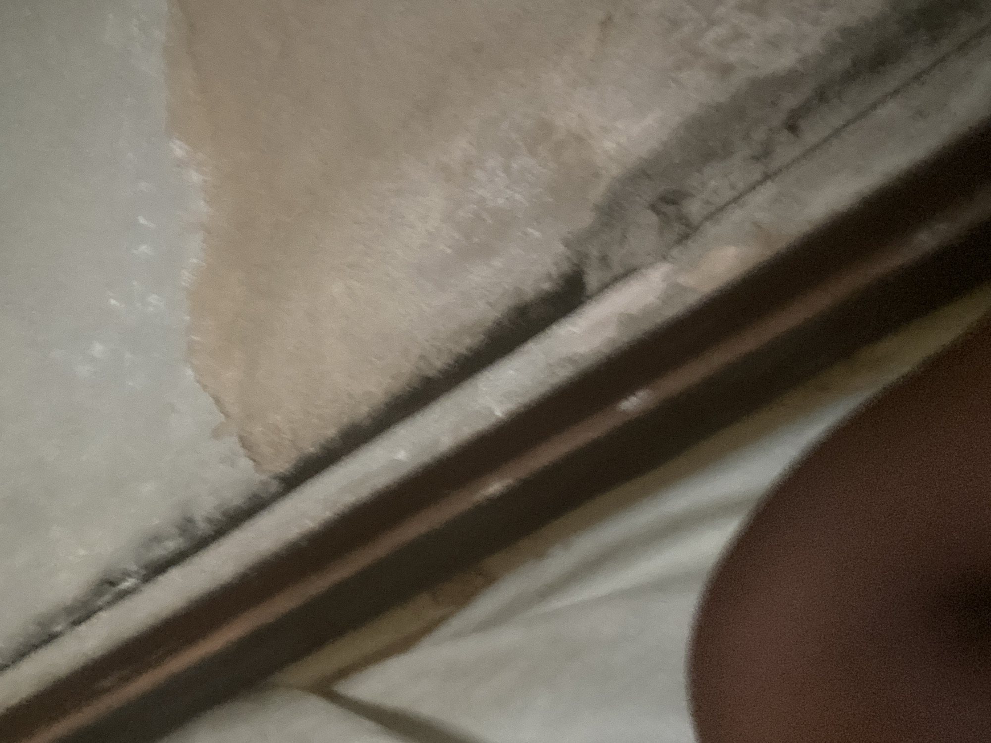 The image shows a close-up of a floor or wall surface with distinct textured areas and linear features. A significant portion of the image displays a rough, sandy-colored surface with patches of lighter and darker grey, possibly indicating water damage or discoloration. Parallel to this textured surface are several dark, linear bands with some lighter brown or copper-colored highlights. The overall impression is one of a weathered or unfinished construction material. In the lower left corner, a rounded, dark brown area is visible, which appears to be part of a person's limb, likely an arm or leg, due to its skin-like texture and curvature. The background, to the left of the linear bands, consists of a softly lit, creased white or off-white surface, resembling fabric like a sheet or curtain. The lighting suggests an indoor environment, possibly dimly lit. Without additional context or clearer details, it is impossible to definitively determine the specific location within Jauro Yinu, Nigeria, or the exact nature of the scene. The image captures a fragment of what could be a domestic setting, a construction site, or an outdoor area with specific architectural features.