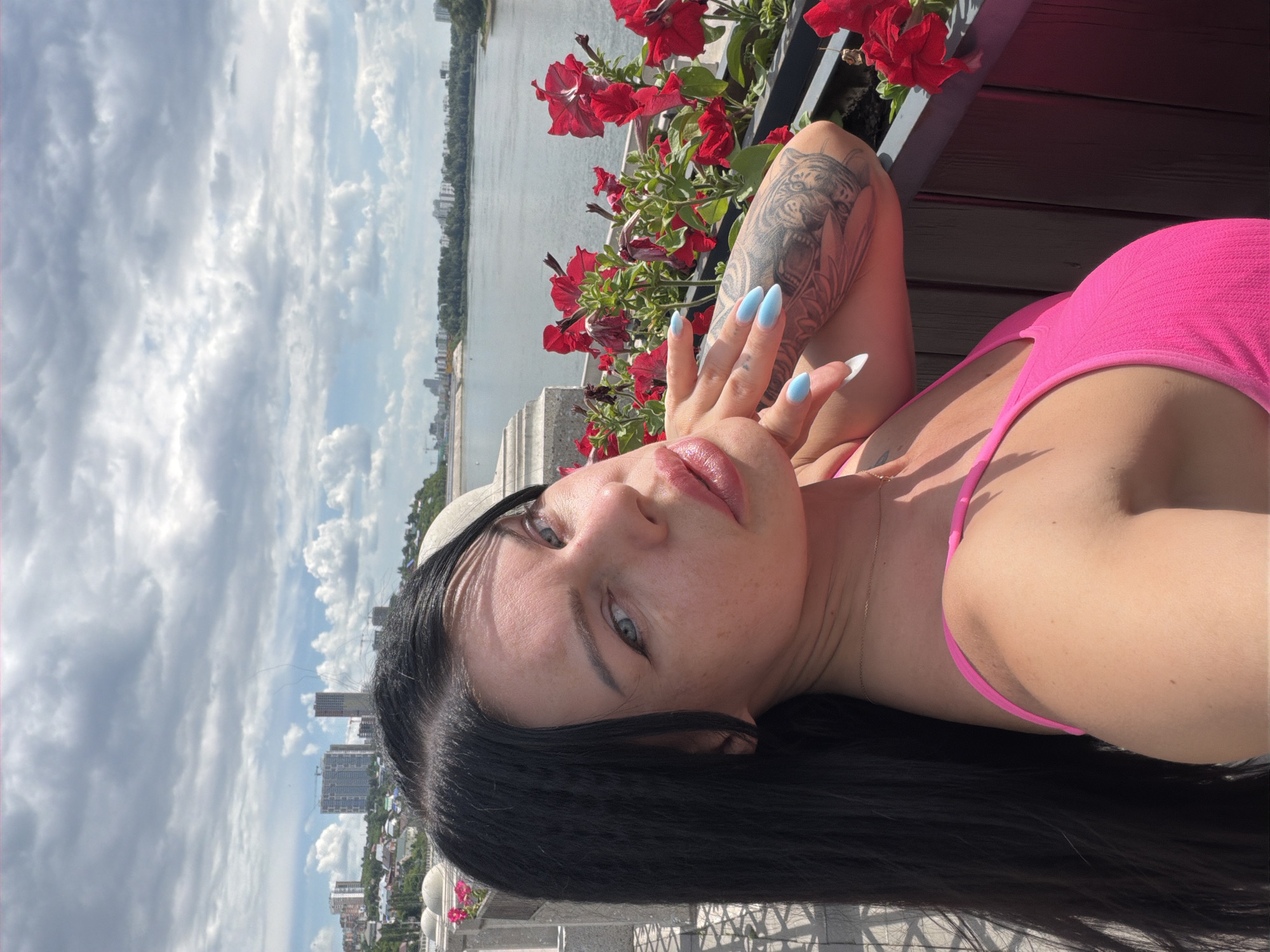 The image shows a woman lying down outdoors with a city skyline and river in the background. She has long, dark hair and is wearing a bright pink top. Her left arm is raised, with her hand near her face. She has light blue painted nails and a tattoo on her forearm. Red flowers are visible in the foreground, suggesting a garden or balcony setting. The sky is cloudy, and the lighting indicates it is daytime, likely late afternoon or early evening due to the long shadows. The woman appears relaxed, with her eyes open and looking towards the camera. The city skyline includes several buildings, some of which appear to be under construction. The presence of a river and urban landscape suggests the location is a city. Based on the provided context, the location is Ufa, Russia.
