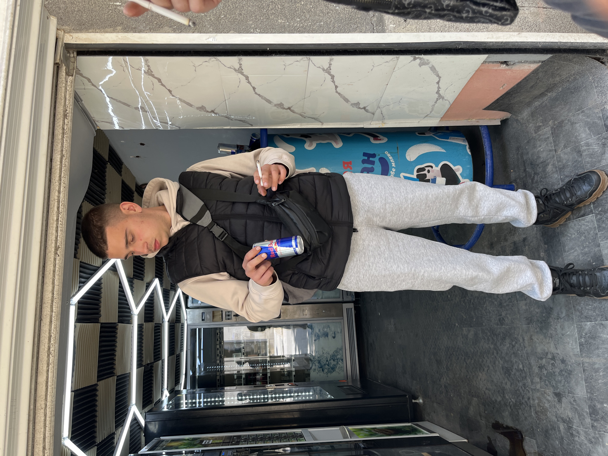 A young man stands in front of vending machines. He is holding a cigarette in his left hand and a can of Red Bull in his right. He wears gray sweatpants, a beige hooded sweatshirt, and a black puffy vest. He also wears a black shoulder bag. The vending machines are situated against a wall with alternating black and white geometric designs, highlighted by vertical neon lights. Behind the man, there is a blue object with Cyrillic script, supported by blue metal bars. The location appears to be an enclosed area, possibly an entrance or vestibule. Above the man are white tiles with marble patterns and some light pink tiles. A hand and the end of a cigarette is visible at the top of the photo. The location in Plovdiv, Bulgaria, cannot be verified without additional information.