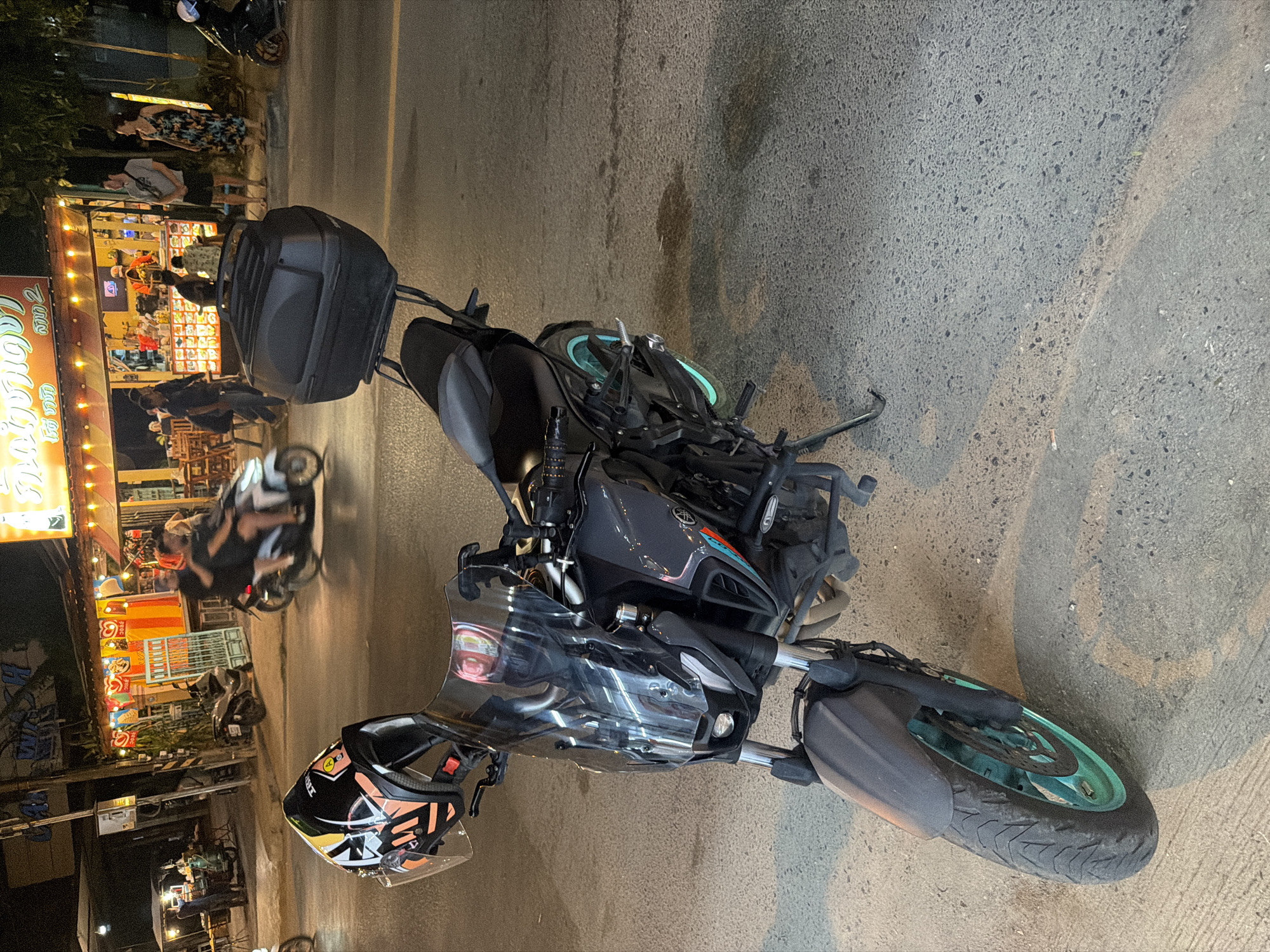 At night on a street in Baan Mae Nam, Thailand, a black motorcycle is parked and angled toward the right of the frame. A black helmet with orange and white accents is attached to the left handlebar. The motorcycle has a black storage box on the back, a tinted windscreen, and teal accents on the front wheel and brake calipers. In the background, a food stall with colorful lights is open. Several people are visible at the food stall, with some interacting and others standing. A few motorcycles are also present on the street. The ground is asphalt.
