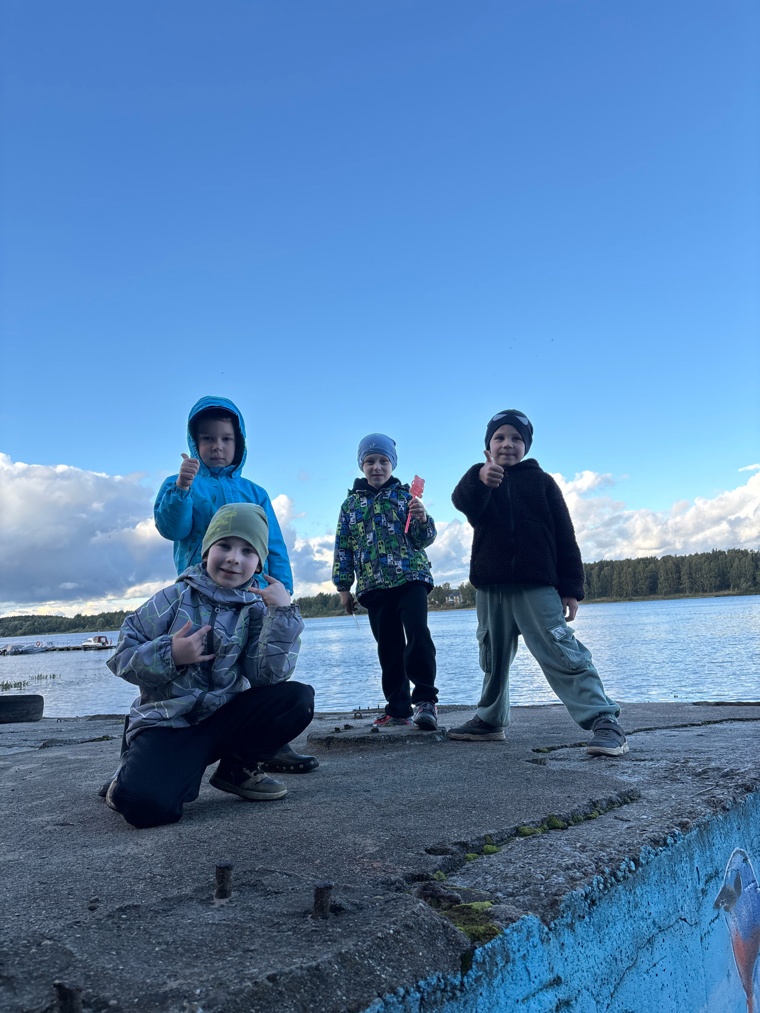 Four young boys stand on a concrete pier by a body of water, likely a lake or river, on an overcast day. The boy on the far left, wearing a blue hooded jacket and a green beanie, gives a thumbs-up. To his right, a boy in a gray and green patterned jacket and green beanie makes a rock-on hand gesture. The third boy from the left, in a colorful jacket with a hood, holds a small pink object. The boy on the far right, wearing a black fleece jacket and light green cargo pants, also gives a thumbs-up. The background shows a treeline, a calm body of water, and a cloudy sky. The concrete pier is weathered and shows signs of disrepair, with some protruding metal rods and mossy patches. There are no visible landmarks that would definitively place this scene in Zhukovo, Russia.