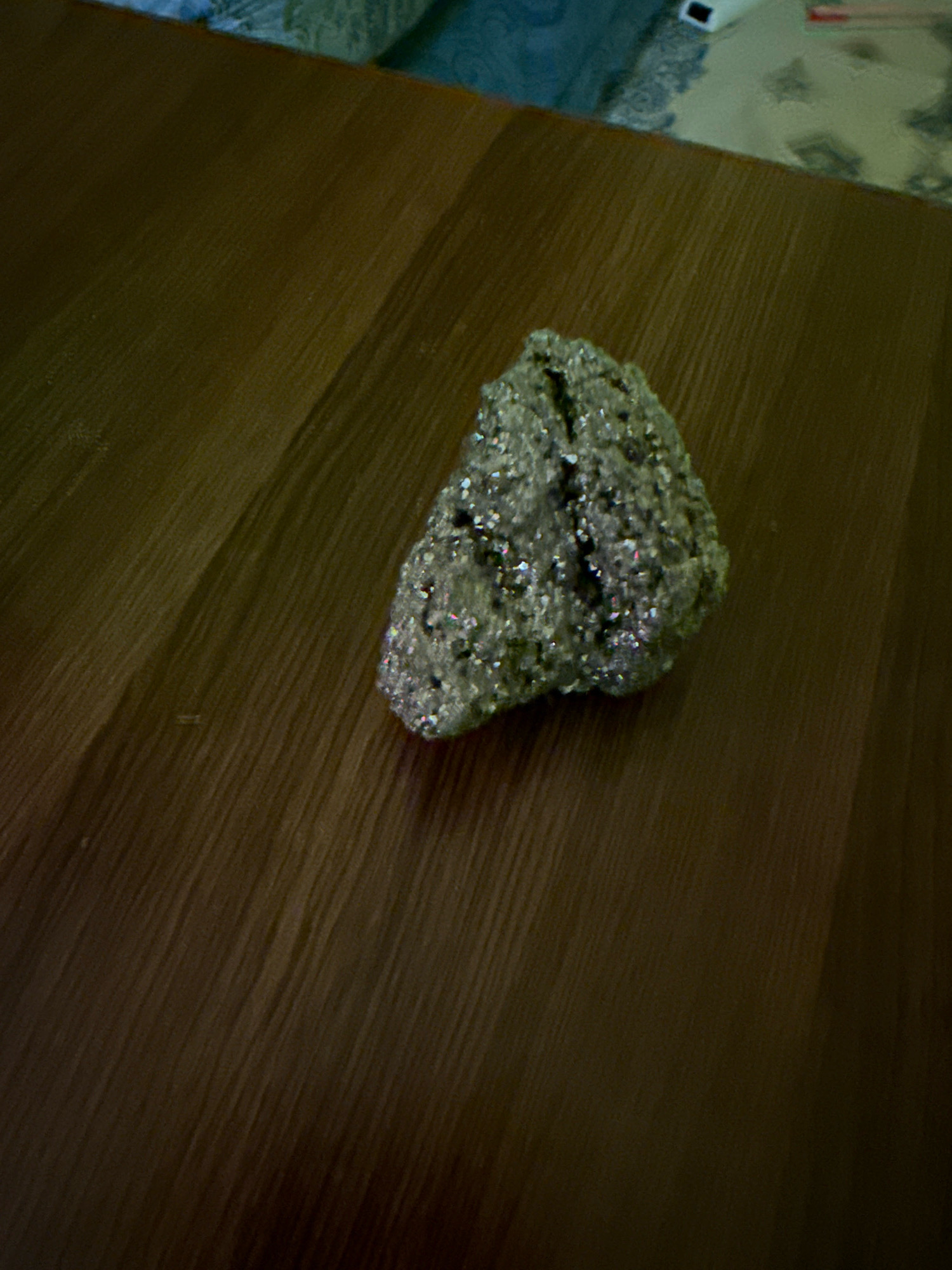 A single piece of pyrite, commonly known as fool's gold, rests on a polished wooden surface. The pyrite is irregular in shape and exhibits a metallic, brass-yellow sheen with some darker, duller areas. The wooden surface has a visible grain pattern running diagonally across the image, suggesting it is a table or countertop. In the upper background, out of focus, are hints of a blue patterned fabric and a white object with red accents. There are no people or animals present in the image, and no contextual clues place the scene specifically in Kolkata, India. The lighting appears to be artificial, casting subtle shadows on the wooden surface.