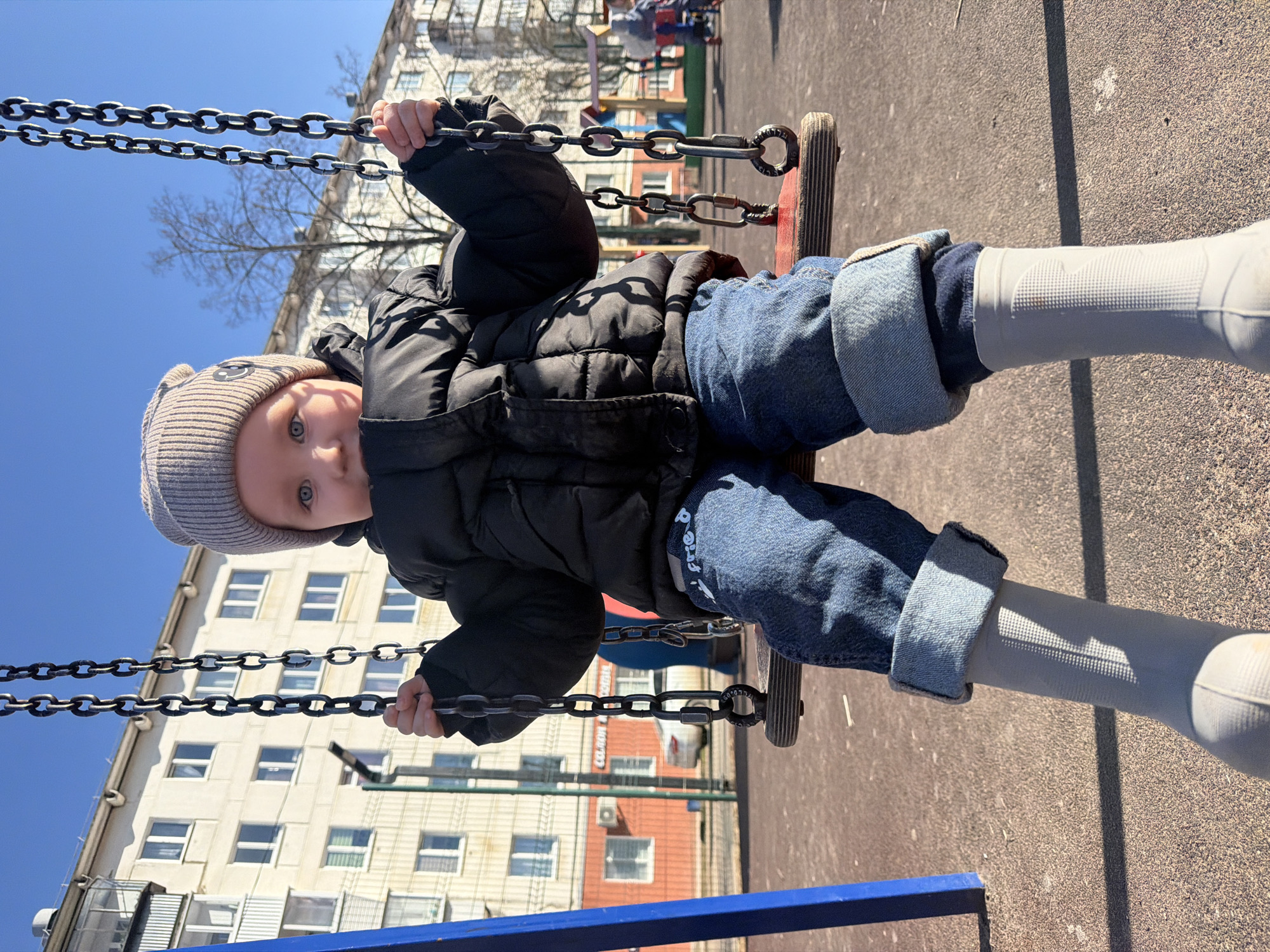 A young child sits on a swing at a playground in Chaikovsky, Russia, on a bright, sunny day. The image is oriented vertically, with the child facing the camera.
The child, likely a toddler or preschooler, is dressed warmly in a black puffer jacket, blue jeans cuffed at the ankles, and light grey or white tall rain boots. They wear a light grey knit beanie with a subtle smiley face design. The child's hands grip the metal chains of the swing, which has a worn wooden seat. Their gaze is directed forward, with a calm and curious expression.
In the background, under a clear blue sky, are large, multi-story apartment buildings, typical of Russian residential architecture. Some buildings have visible windows and balconies. A bare tree, indicating early spring or late autumn, stands between the child and the buildings. The ground beneath the swing is paved with asphalt or a similar material, and strong shadows are cast by the swing chains and the child, suggesting bright daylight, possibly midday or early afternoon. The overall scene depicts a quiet moment at a playground on a cool, clear day.
No clearly legible text is visible, though a faint logo or text element might be present on the child's jeans, it is indistinct.