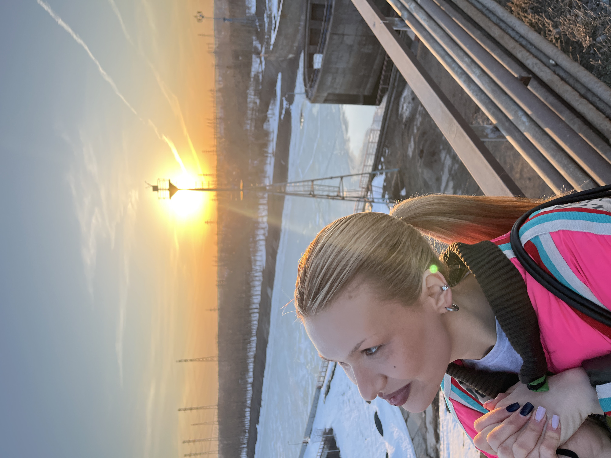 A young woman with blonde hair pulled back into a ponytail, wearing a pink jacket, stands on an elevated platform in Chaikovsky, Russia. She is looking down at a frozen body of water, possibly the Kama River. In the background, a bright sunset illuminates the cityscape with a golden glow, revealing power lines and industrial structures. The platform is constructed with metal pipes and beams. The woman's hands are clasped together, and she appears to be observing the scene below.