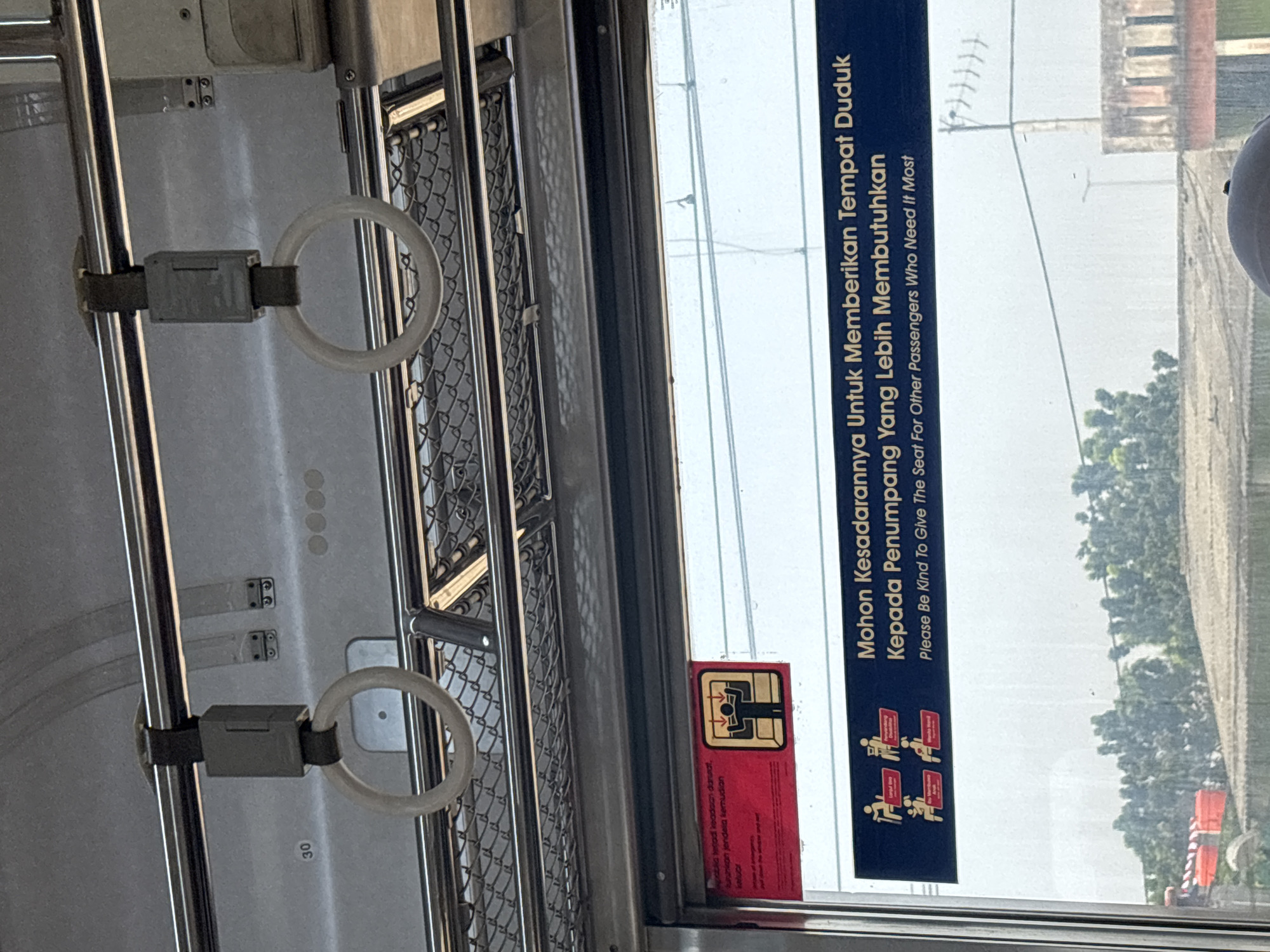 The image shows the interior of a public transportation vehicle, likely a train or bus, in North Jakarta, Indonesia. In the foreground, there are two white ring-shaped handholds hanging from a metal bar above. Below the handholds, a window reveals a blurred outdoor scene with trees and buildings. A sign is affixed to the window, with a red section at the top and a blue section below. The red section contains an illustration of a person with arms outstretched. The blue section has text in Indonesian and English, indicating a request to give up seats to passengers who need them more, featuring icons of various individuals like the elderly, pregnant women, and those with children. The overall impression is of a public transport environment, likely during daytime given the lighting.
