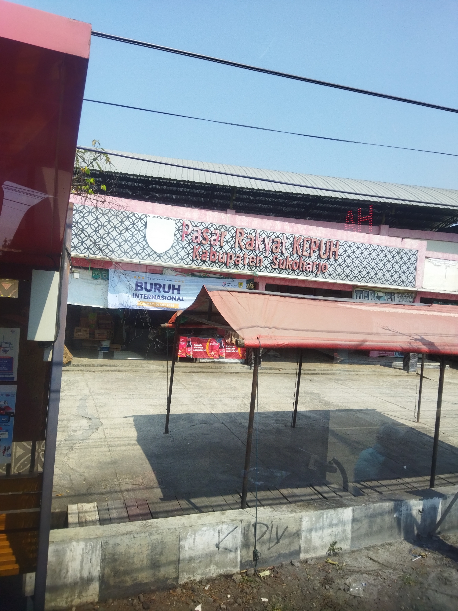 The image captures a street-level view of a market building, likely in Begajah, Indonesia, on a clear, sunny day. The building features a pink facade adorned with a white shield emblem and a repeating black circular pattern. Above the entrance, large red lettering spells out "Pasar Rakyat Kepuh Kabupaten Sukoharjo," indicating it's the Kepuh People's Market in Sukoharjo Regency. A banner is hung across the building, reading "SELAMAT HARI BURUH INTERNASIONAL" with "MAI, 01 MEI 2019" below it, signifying International Labor Day on May 1, 2019. Several stalls or structures with red canvas awnings are set up in front of the market building, some with merchandise displayed. The ground in front of the market is paved, with some areas showing wear and tear, and a concrete barrier runs along the foreground. Overhead, power lines are visible against the bright blue sky. The scene appears to be an outdoor commercial area, possibly with some seating areas or waiting spots.