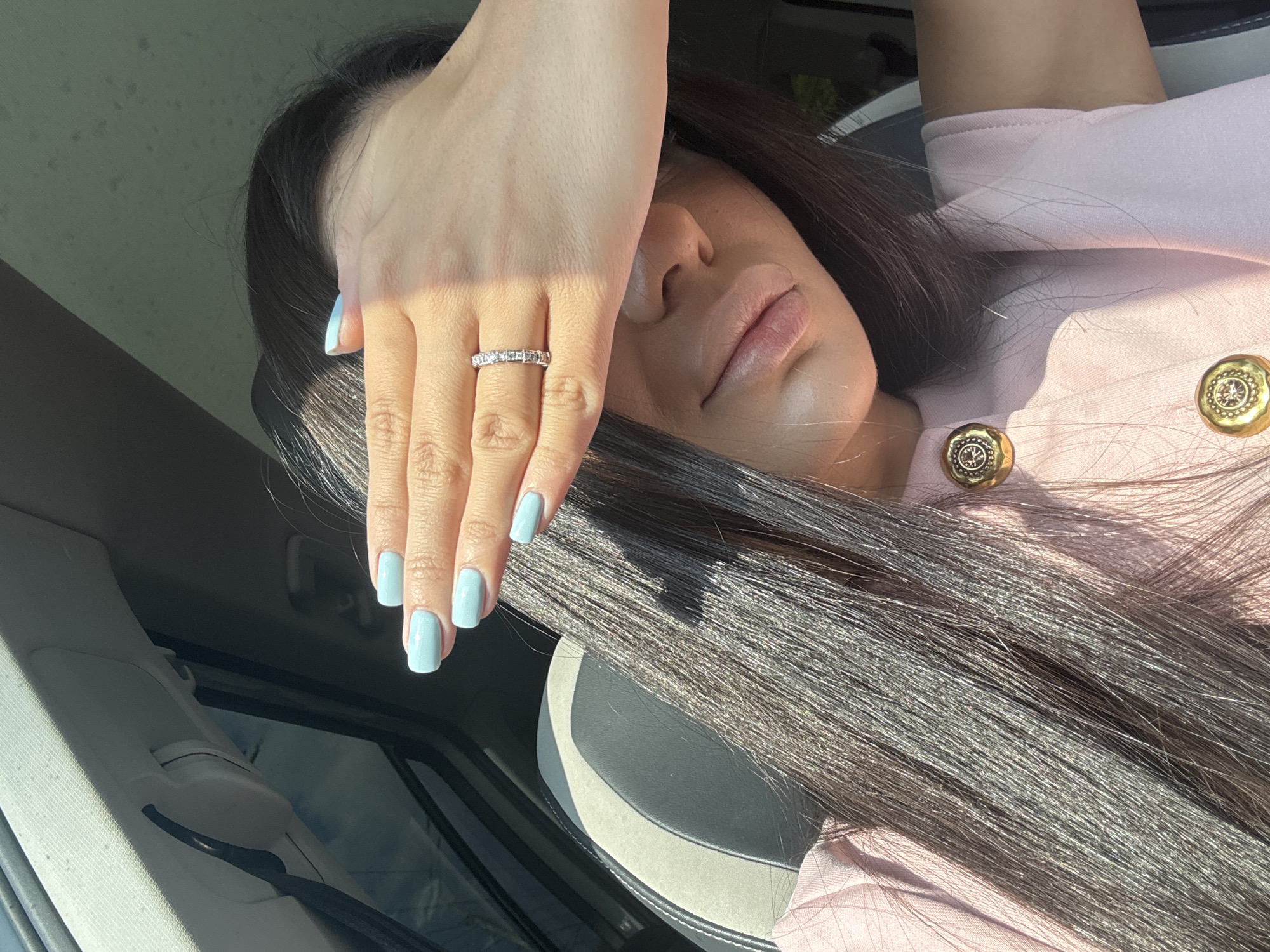 The image depicts a woman in a car. She is fair-skinned with long, dark hair, light blue manicured nails, and is wearing a diamond ring. She is wearing a light pink shirt with decorative gold buttons. Her hand is shielding her face from the sun. The car interior is visible, including the seat and part of the door. The setting is likely Yerevan, Armenia based on the user's request, although no visual elements directly confirm this location.