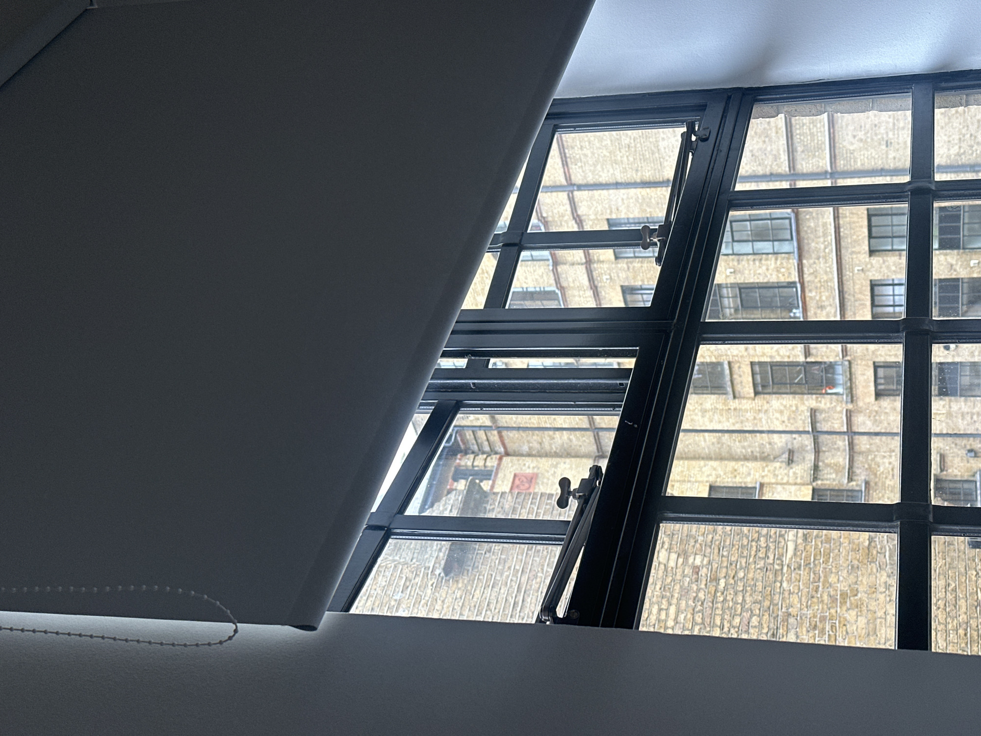 The image shows a partially opened window with black metal frames looking out onto a brick building. The building exterior has a brick facade with multiple window units and metallic bars. A light-colored roller blind is partially obscuring the view from the left side of the frame. The scene is indoors and could be located in London, United Kingdom, due to the architecture and window styles that are popular in the city.
