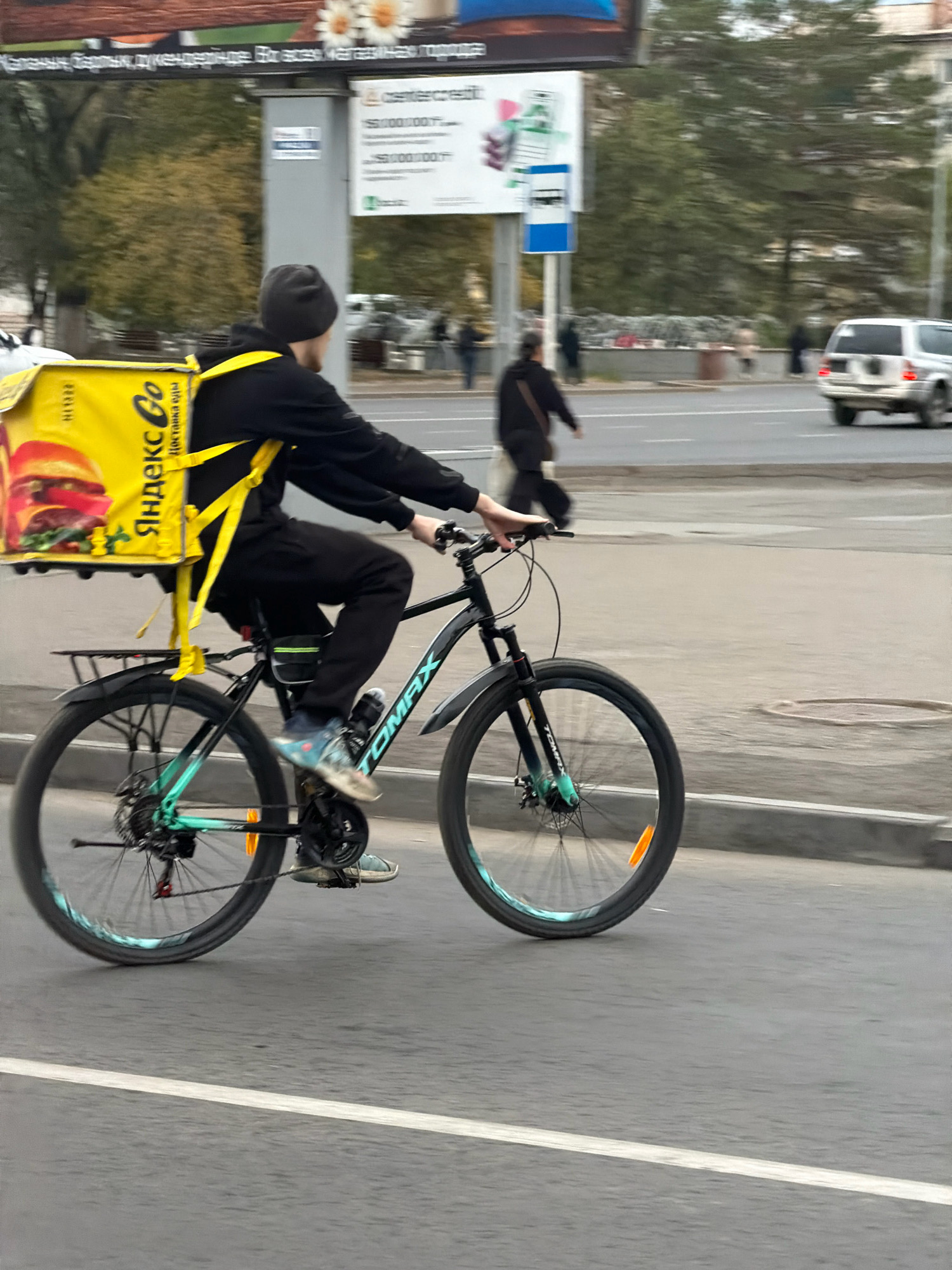 A delivery person on a bicycle is captured in motion in the city of Pavlodar, Kazakhstan. The person, wearing a dark beanie and black clothing, is riding a black bicycle with teal accents. Strapped to their back is a large, bright yellow delivery box with the Yandex.Go logo and an image of a burger. The delivery person's left foot is on the pedal, indicating they are actively riding. In the background, other individuals are visible walking and a white SUV is driving on the road. The scene appears to be a street in an urban environment, with buildings and trees lining the background. The image is slightly blurred due to the motion of the bicycle and possibly the camera.