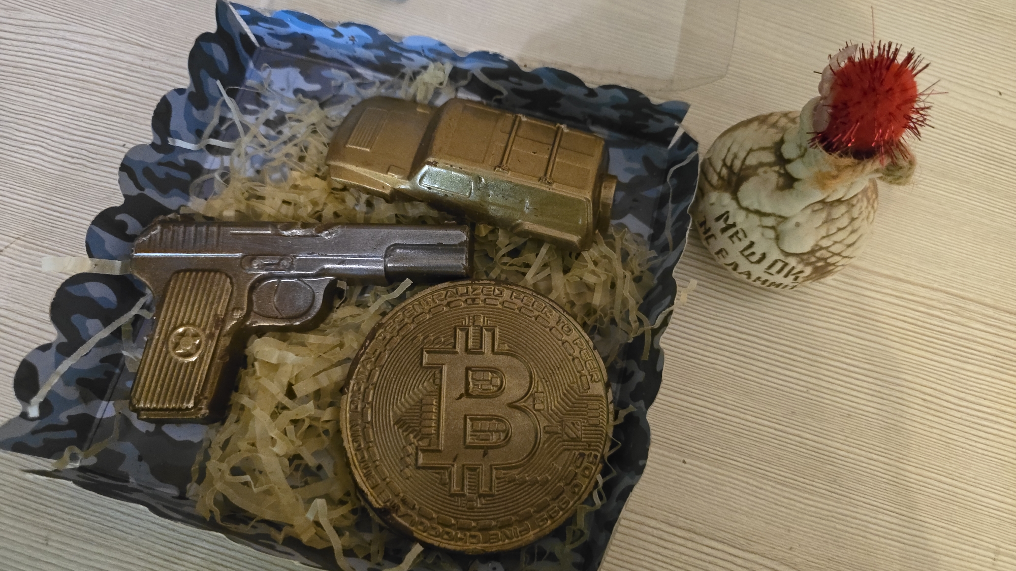 A close-up, top-down view of a gift box with a camouflage interior on a wooden surface. The box contains several chocolate replicas: a Bitcoin coin, a handgun, and a magazine. To the upper left of the box, a small, round, light-colored object with a red, spiky pom-pom on top is visible. The object has some inscription in what appears to be Cyrillic script. The wooden surface is light brown with distinct grain lines. The overall lighting suggests an indoor setting. The location of Качинский округ, Ukraine is not visible in the image.
