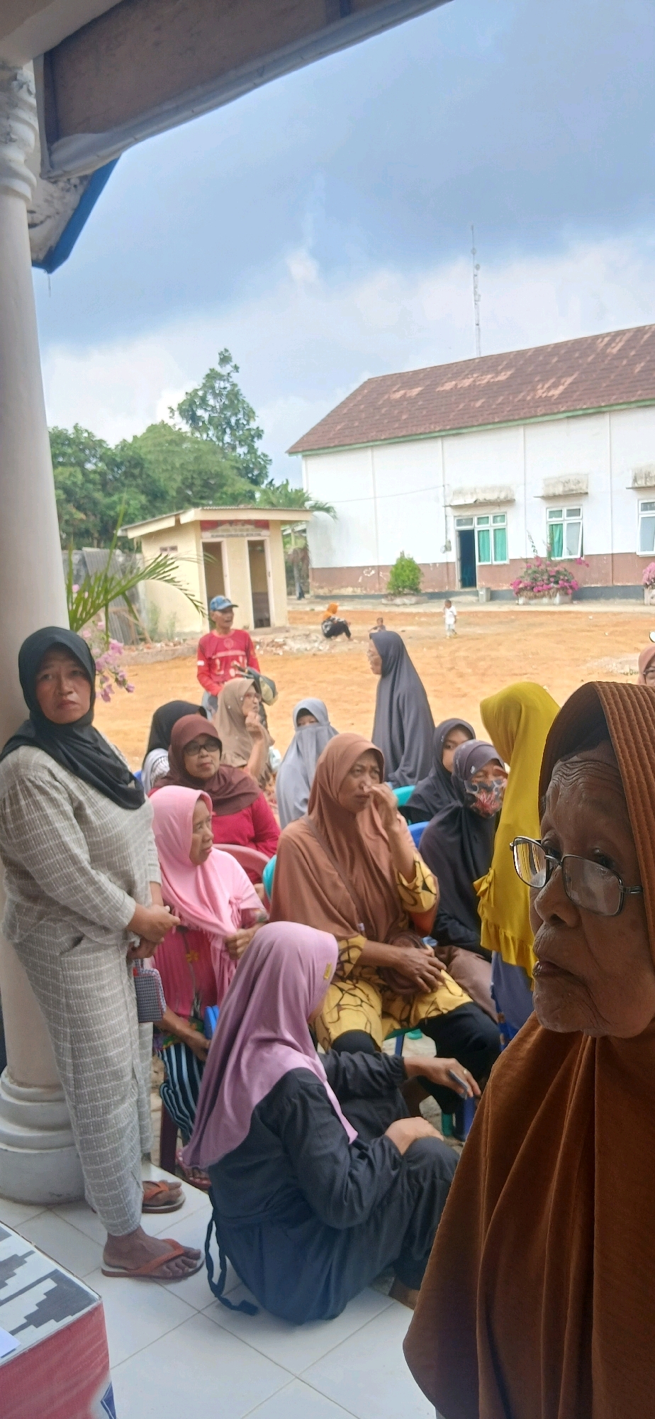 The image shows a group of people, mostly women wearing hijabs, gathered outdoors. They appear to be waiting or attending an event. In the background, there is a building with white walls and a brown roof, and some trees. The ground is sandy and there are a few small structures. The sky is overcast. The people are dressed in casual clothing. The scene suggests a community gathering or an outdoor event in a rural or semi-rural setting.