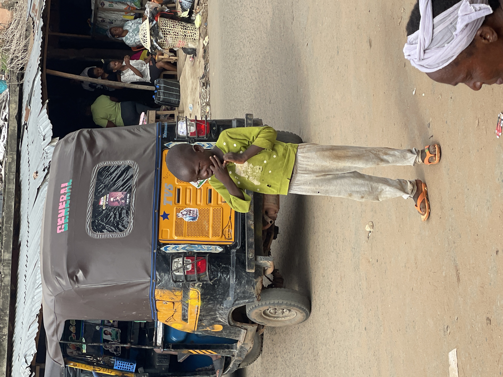 In Nkpor, Nigeria, a young Black boy wearing a light green long-sleeved shirt with white speckles, light gray pants, and orange sandals stands in front of a parked yellow and black "TVS General" auto rickshaw, touching his face. To the left of the rickshaw is a lean-to structure with corrugated metal roofing, where people are sitting. To the right of the boy, a darker-skinned Black man with a white head covering is partially visible. The ground appears to be a paved road.