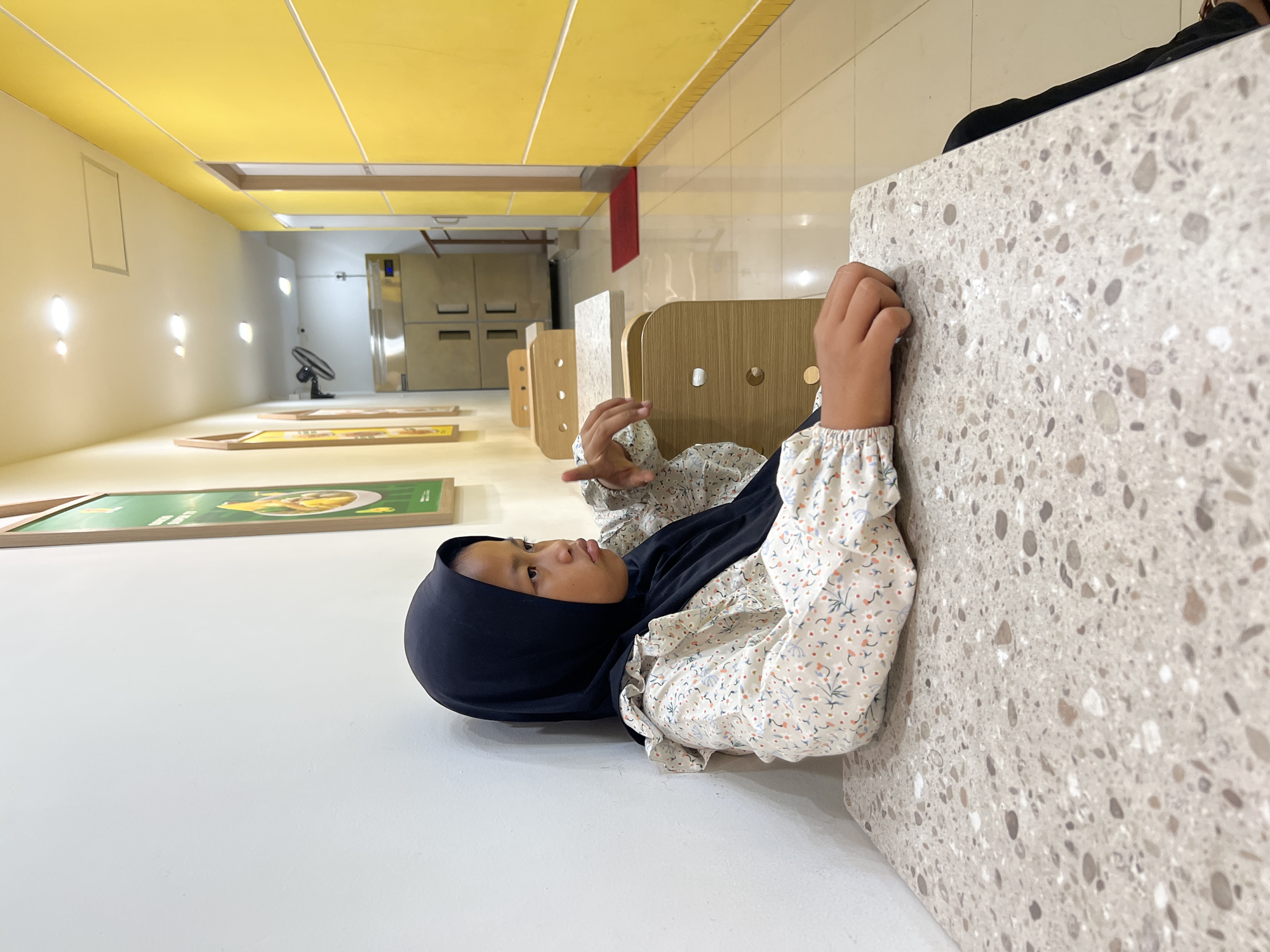 In Gunung Putri, Indonesia, a young girl wearing a black hijab and a floral-patterned shirt sits at a speckled grey table in a brightly lit hallway with yellow walls. She is facing left, with her elbows resting on the table and her hands clasped. Her expression is thoughtful as she gazes off to the side. Behind her, there are framed pictures on the wall, and further down the hallway, kitchen equipment and doors are visible.