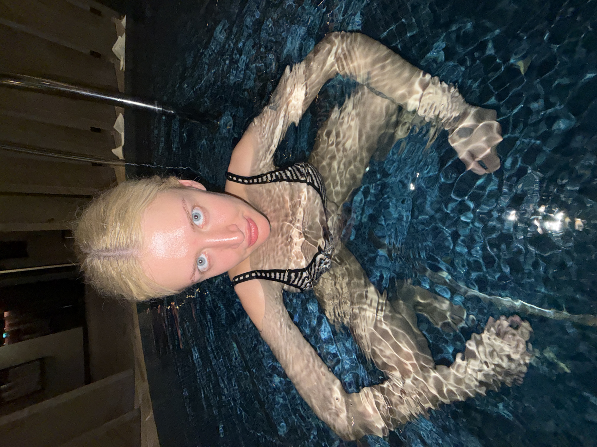 A woman with blonde hair and blue eyes is in a pool of water. She is wearing a black and white bikini top. The pool water is blue and reflects light. The woman is looking at the camera. A metal bar is visible in the upper left corner of the image. Parts of a wall can be seen in the background. The location appears to be an indoor pool, but there is not enough information to determine whether it is in Vracar Urban Municipality, Serbia.