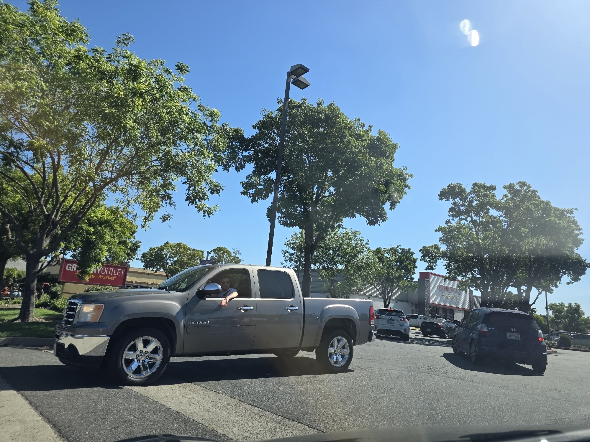 The image captures a sunny day scene in West Sacramento, USA, likely near a commercial area. In the foreground, a grey pickup truck is driving on a paved road, with a driver visible inside wearing sunglasses. The truck has chrome wheels and a short bed. Behind the truck, several other vehicles are present, including a dark-colored hatchback with a license plate starting with "7MT". Further in the background, a building with a red and white sign that reads "GROCERY OUTLET" is visible, surrounded by trees and a street light. The sky is clear and blue.