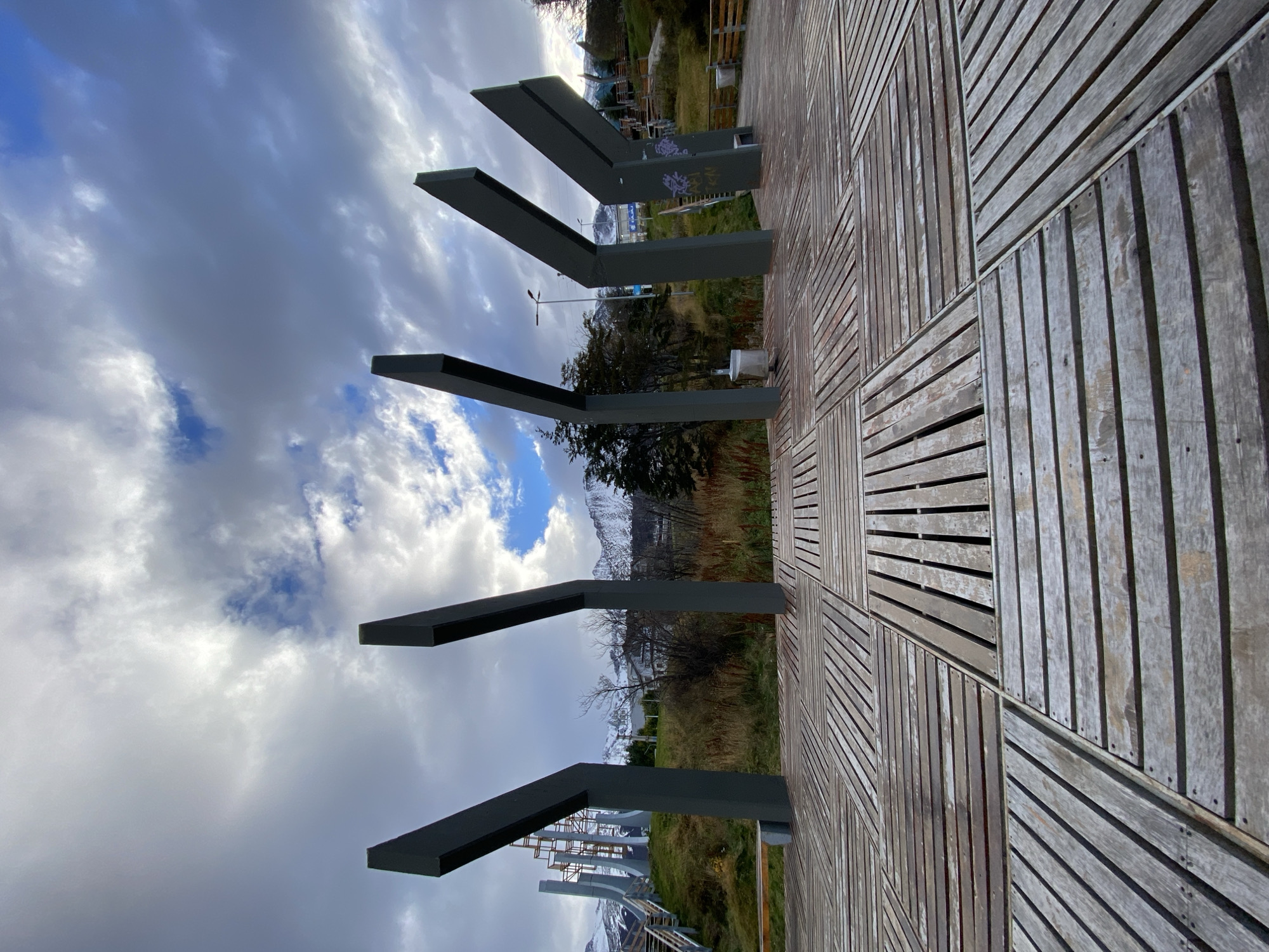 The image shows a perspective shot from a wooden boardwalk in Ushuaia, Argentina. The boardwalk features a distinctive pattern of alternating rectangular sections of wooden planks arranged perpendicularly. Metal structures resembling the letter "L" are mounted on the boardwalk. The background reveals a landscape with grass, trees, and mountains capped with snow in the distance. Overhead, the sky is filled with clouds.
