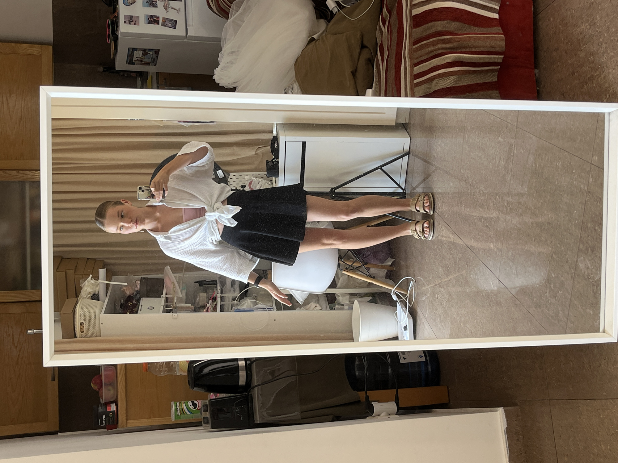 A young woman stands in a room, taking a mirror selfie. She is wearing a white, unbuttoned shirt tied at the waist, a pink tank top, and a black skirt. She has on beige sandals and her hair is pulled back. The room appears to be a bedroom or living area, with a bed visible in the reflection. The environment is cluttered with various items, including shelves stocked with personal belongings, a refrigerator, and furniture. The floor is tiled. The room has light-colored curtains in the background. There is no clear indication of the specific location in Budva, Montenegro, beyond the general appearance of an indoor residential setting.