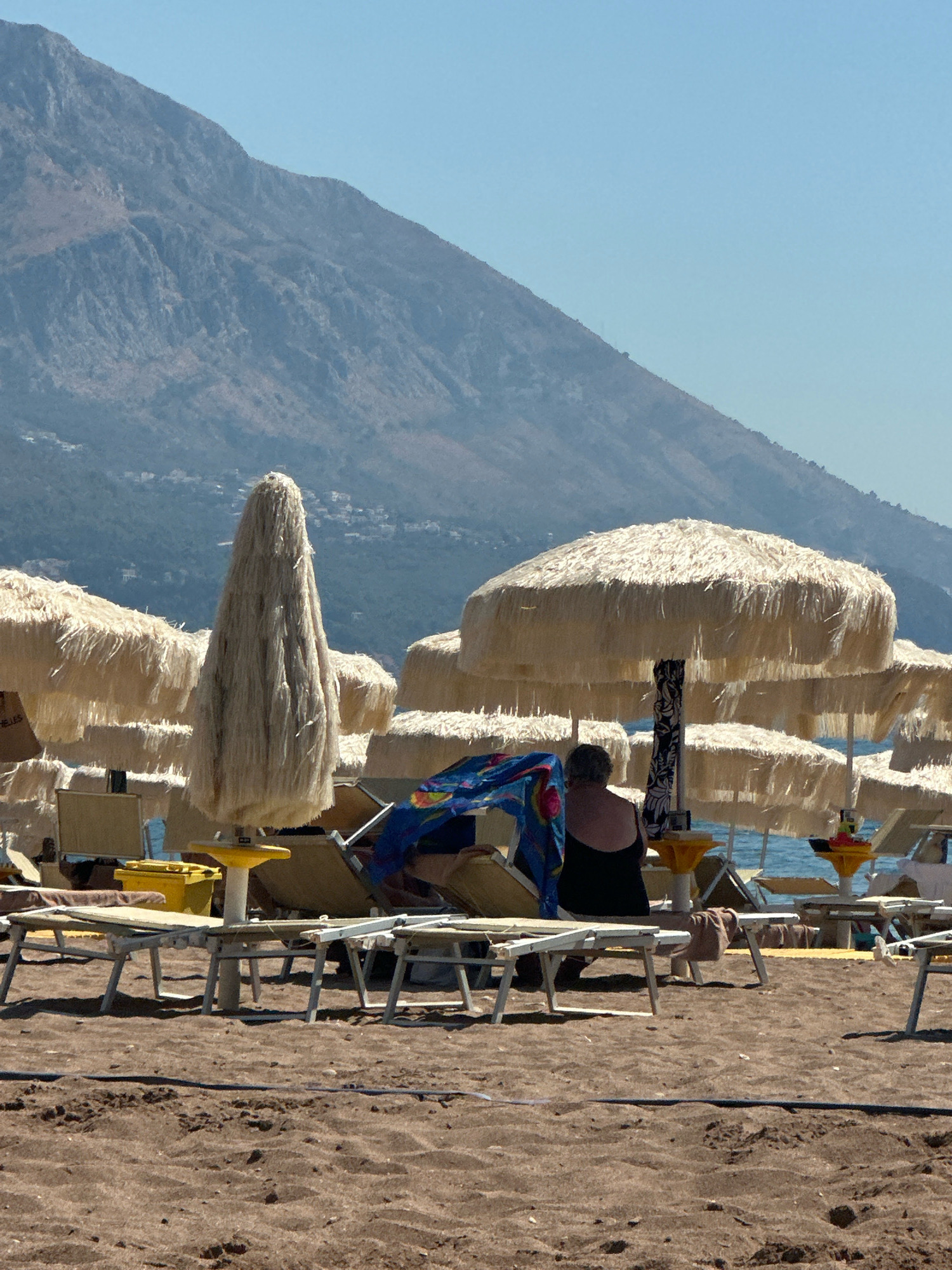The image depicts a beach scene in Boreti, Montenegro, with a mountainous backdrop under a clear blue sky. In the foreground, several beach chairs and umbrellas are set up on the sandy shore. One person, wearing a black bathing suit, is seated on a lounge chair. The umbrellas are made of straw-like material. The sea is visible in the background.