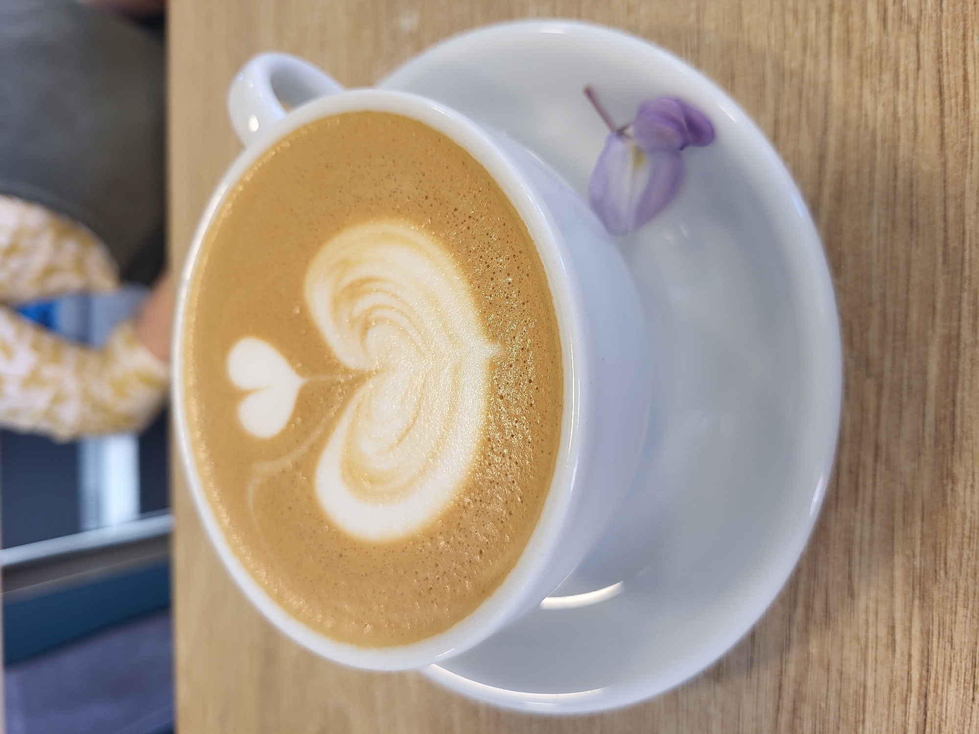 A top-down view shows a white coffee cup with latte art in the shape of a heart, resting on a white saucer. A small purple flower lies on the saucer next to the cup. The cup and saucer are placed on a light brown wooden table. Part of a person is visible in the left side of the frame, wearing a patterned yellow and white shirt, sitting on what appears to be a dark grey chair. The location is likely a cafe in Budva, Montenegro.