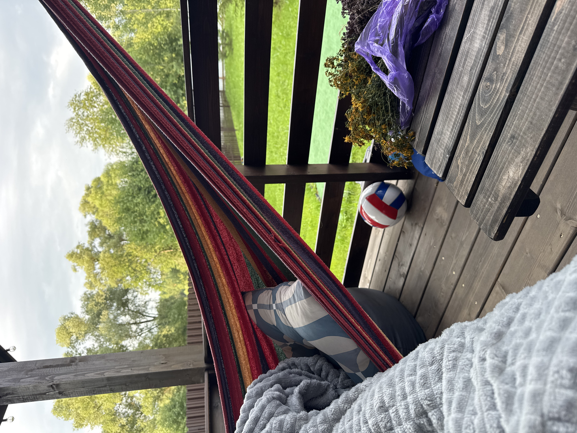 The image depicts a relaxed scene on a wooden deck, presumably in Gavshino, Russia. A colorful striped hammock is stretched across the frame, partially occupied by a person covered with a grey, textured blanket and a pillow with a blue and white geometric pattern. Next to the hammock, on the wooden floor, rests a red, white, and blue ball. To the right, a low wooden table holds a bunch of dried yellow and green herbs tied with a purple plastic bag. The background reveals a lush green lawn bordered by a dark wooden fence, with trees and a cloudy sky visible beyond. The overall impression is one of a quiet, outdoor leisure space.