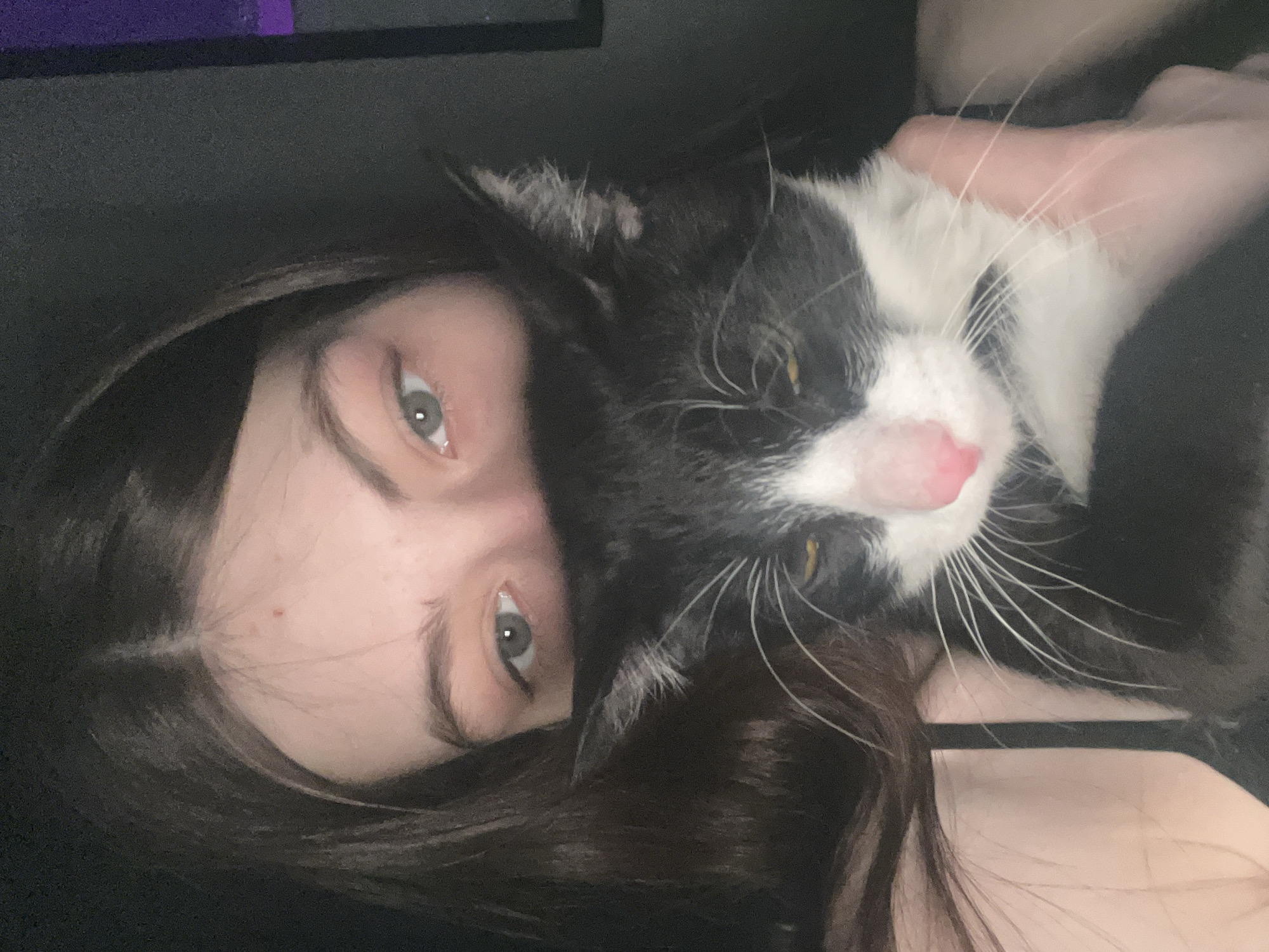 A selfie-style photograph features a young woman with long, dark hair and light eyes. She is tilted to the side, with her face partially obscured by a black and white cat she is holding close to her. The cat has a pink nose and long white whiskers. A hand is visible holding the cat. The background is dark and indistinct. The location is not discernible from the image.
