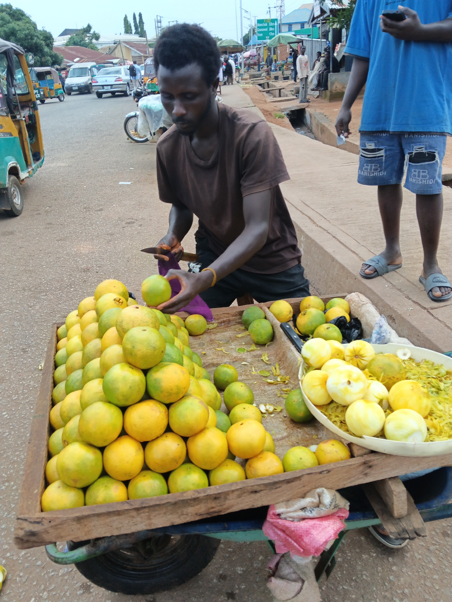 In an outdoor market scene, a dark-skinned man with short, dark hair stands behind a wooden cart piled high with oranges. He wears a brown t-shirt and dark shorts, and a yellow bracelet adorns his left wrist. In his right hand, he holds a small knife and is in the process of slicing a green orange. Another man, wearing a blue t-shirt and denim shorts, stands to the right, holding a smartphone. The cart is positioned on a paved surface next to a raised concrete walkway. Beyond the immediate scene, a yellow and green motorized tricycle is parked to the left. In the background, other vehicles, market stalls with awnings, and people are visible, suggesting a bustling urban environment.
