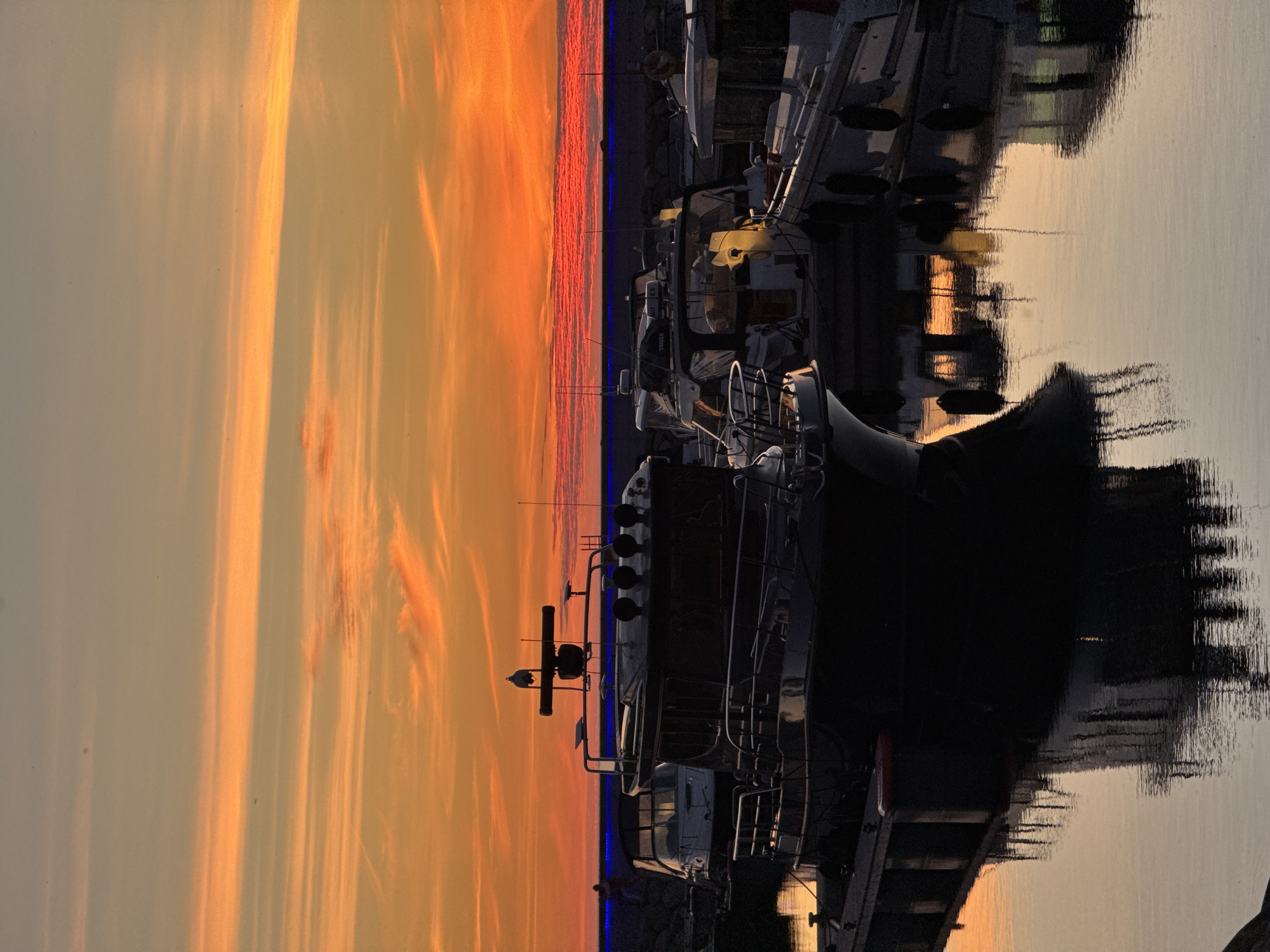 At dusk, a marina in Tallinn, Estonia, is bathed in the warm glow of an orange and yellow sunset. The sky is streaked with clouds, painted with vibrant hues of orange and red, transitioning to a pale blue at the top. Several boats are docked, their dark silhouettes contrasting with the colorful sky and its reflection on the calm water. The foreground features the stern of a dark-hulled motor yacht, its railings and windows visible. Behind it, other boats are moored, including a white motor yacht and what appears to be a ferry or larger vessel in the background. A rocky breakwater runs along the left side of the frame, and a single figure can be seen standing near it. Blue lights are visible along the breakwater. The water mirrors the sky and the boats, creating a serene and tranquil atmosphere.