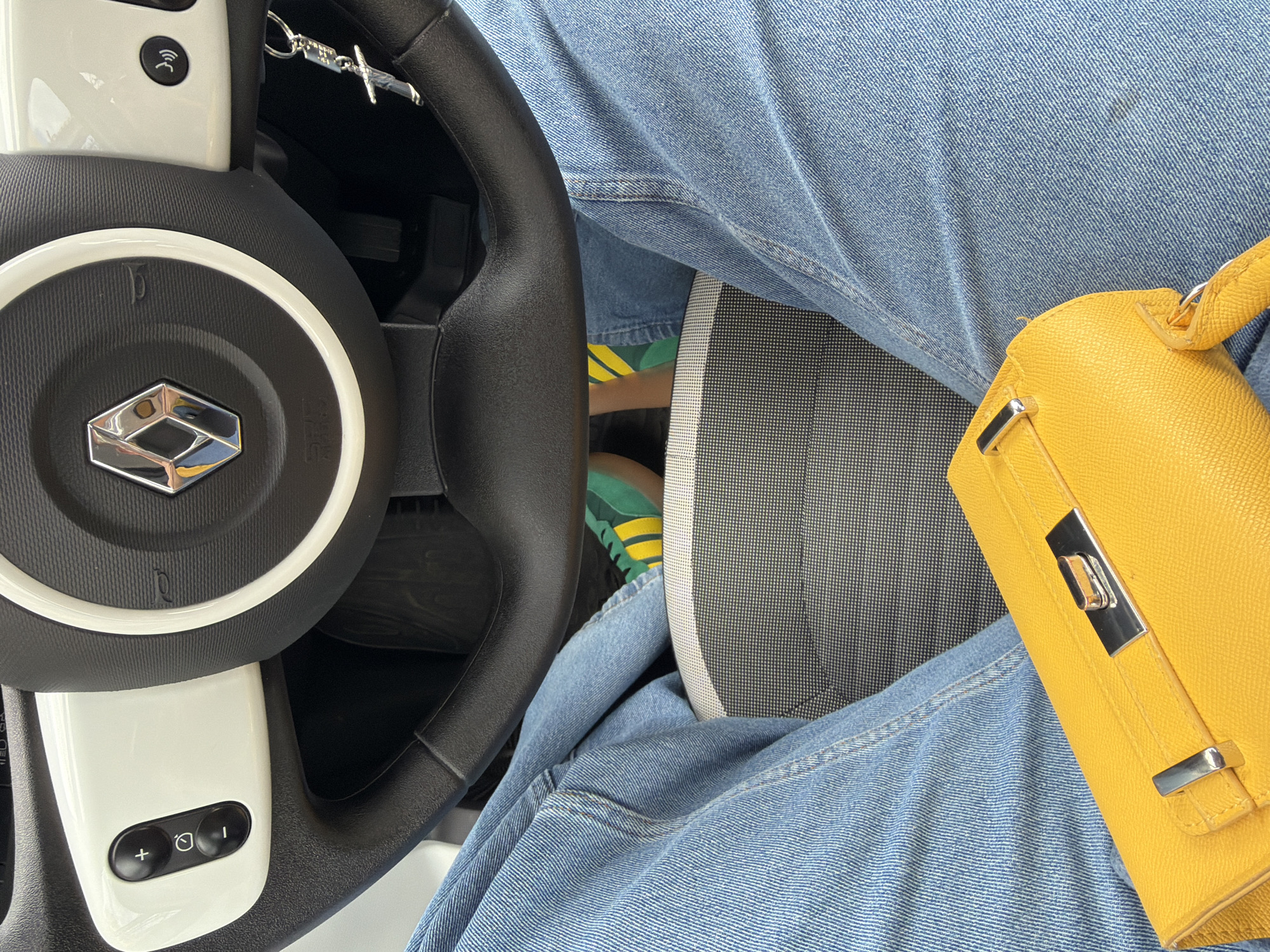 A view from inside a car, showing the driver's perspective, presumably in Buhovo, Bulgaria, although there are no visible location markers in the frame. The driver is wearing light-wash denim jeans and green and yellow athletic shoes. A yellow leather handbag rests on their lap. The car's steering wheel features a logo in the center and controls on the white spokes. A keychain hangs from the ignition. The car seat has a black and white patterned design.