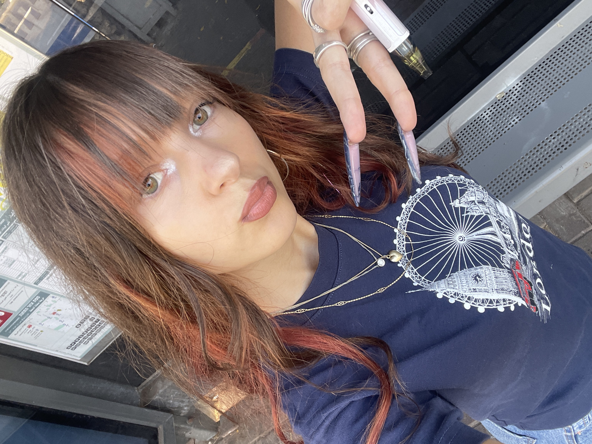 A young woman with long brown hair and auburn highlights is taking a selfie. She is wearing a dark blue t-shirt with a graphic of the London Eye and Big Ben. She has on multiple rings on her fingers and long, pointed fingernails with a marbled design. She is holding a white and pink pen-like object in her right hand. The background appears to be an outdoor setting, possibly a bus stop, with a public information board visible behind her. The lighting suggests it is daytime. The location is Raanana, Israel.