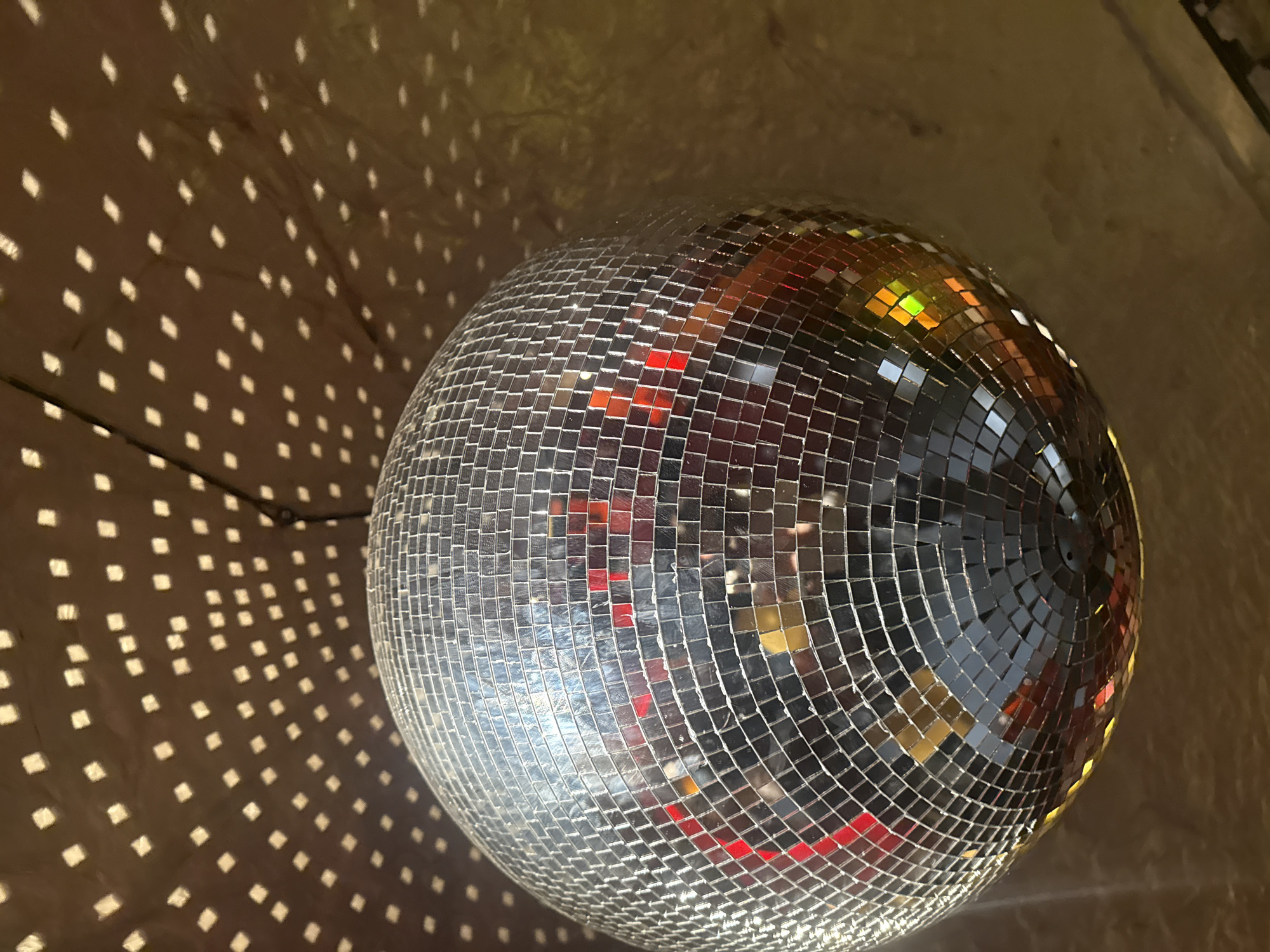 A disco ball hangs from a ceiling, casting a pattern of reflected light onto a wall. The disco ball is silver and covered in small square mirrors. The reflected light pattern is composed of many small, square shapes. A dark cable is visible near the top left corner of the image. The location is Yerevan, Armenia.