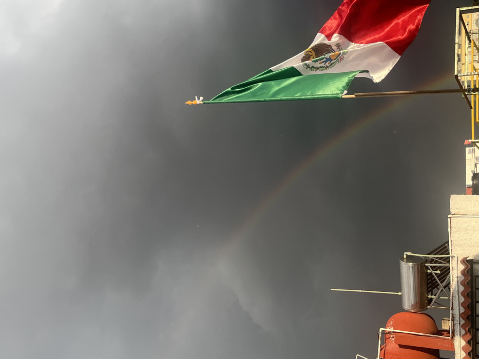 The image presents an outdoor scene dominated by a Mexican flag against a dark, cloudy sky with a rainbow.
**Location:** Unknown city, Unknown state, Mexico.
**Environment:** The sky is filled with dense, dark grey storm clouds, suggesting overcast or post-rain conditions. A faint rainbow arc is visible, extending from the middle-left of the frame towards the upper-right. The foreground appears to be a building rooftop.
**Subjects/Objects:**
* A Mexican flag, featuring green, white (with the national coat of arms depicting an eagle on a nopal devouring a snake), and red vertical stripes, is prominently displayed on a pole on the right side of the frame. The flag is partially unfurled, indicating a light breeze. The flag pole is topped with a distinct yellow finial.
* In the lower-left foreground, several rooftop elements are visible: a large red cylindrical water tank, and a silver horizontal cylindrical solar water heater mounted on a frame, complete with white PVC piping. A section of a white corrugated roof and a white parapet wall are also present.
* No people are visible in the scene.
**Actions/Interactions:** The flag is caught in a gentle breeze, causing it to unfurl. The combination of dark clouds and a rainbow indicates that sunlight is breaking through after a recent period of rain.