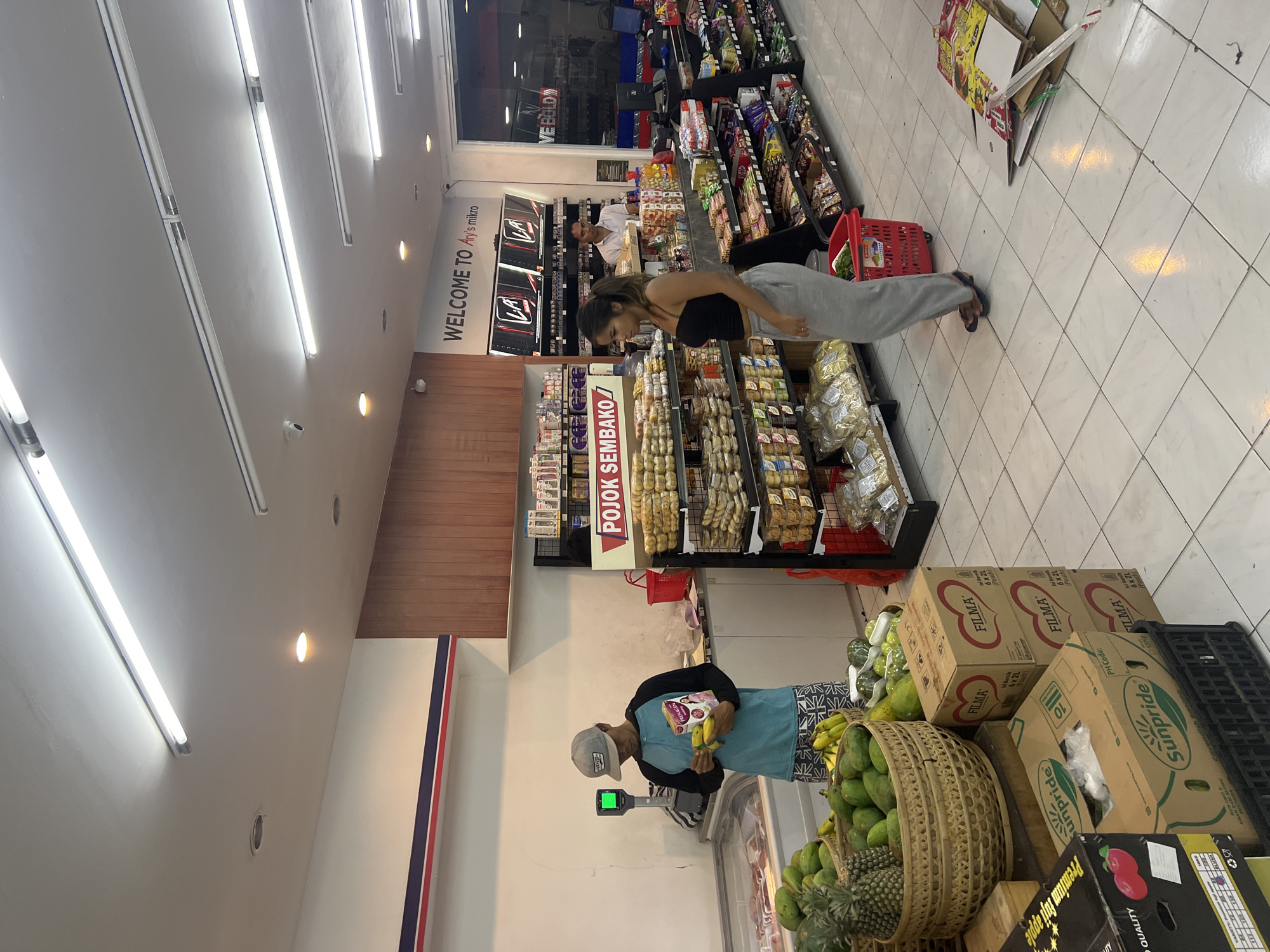 Here is a detailed description of the image:
**Scene:** The image depicts the interior of a small supermarket or convenience store, likely in Sukawati, Indonesia, as indicated by the text “WELCOME TO Ary’s mikro” near the ceiling.
**Environment:** The store has bright fluorescent lighting and tiled floors. The shelves are stocked with various products, including snacks, pastries, and fresh produce. Signage in Indonesian is visible.
**People:**
* A woman is standing in front of a shelf looking at the products. She is wearing a black top and grey sweatpants.
* A man wearing a blue t-shirt, patterned shorts, and a baseball cap is standing near a scale and weighing bananas.
**Objects:**
* Various food products on shelves and in baskets (snacks, pastries, fruits).
* A red shopping basket.
* Cardboard boxes of produce, labeled with brands such as “FILMA” and "Sunpride".
* A scale for weighing produce.
**Actions:**
* The woman is browsing or selecting items on a shelf.
* The man is weighing bananas on a scale, presumably preparing to purchase them.
**Interactions:** The woman is focused on the products on the shelf, and the man is occupied with weighing the bananas. There doesn't appear to be any interaction between them or with any employees.