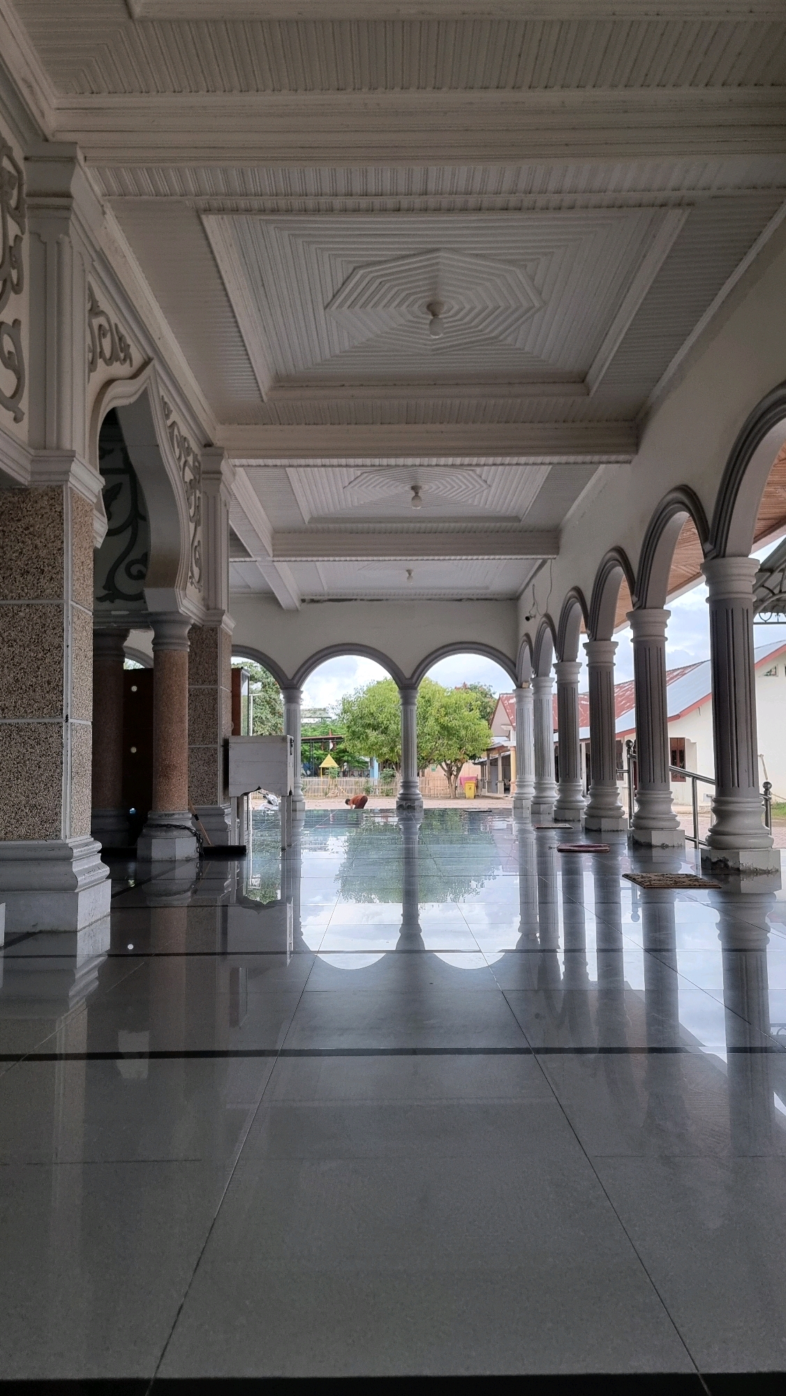The image depicts the exterior walkway of a mosque in Pantegurah, Indonesia. The walkway features a series of arched colonnades and is paved with polished, reflective tiles that mirror the architecture and the sky. Large, ornate pillars support the structure. In the background, through the arches, trees, houses, and a playground are visible. One person is seen kneeling on the ground outside, facing away from the camera. The ceiling of the walkway has intricate geometric paneling and decorative light fixtures.