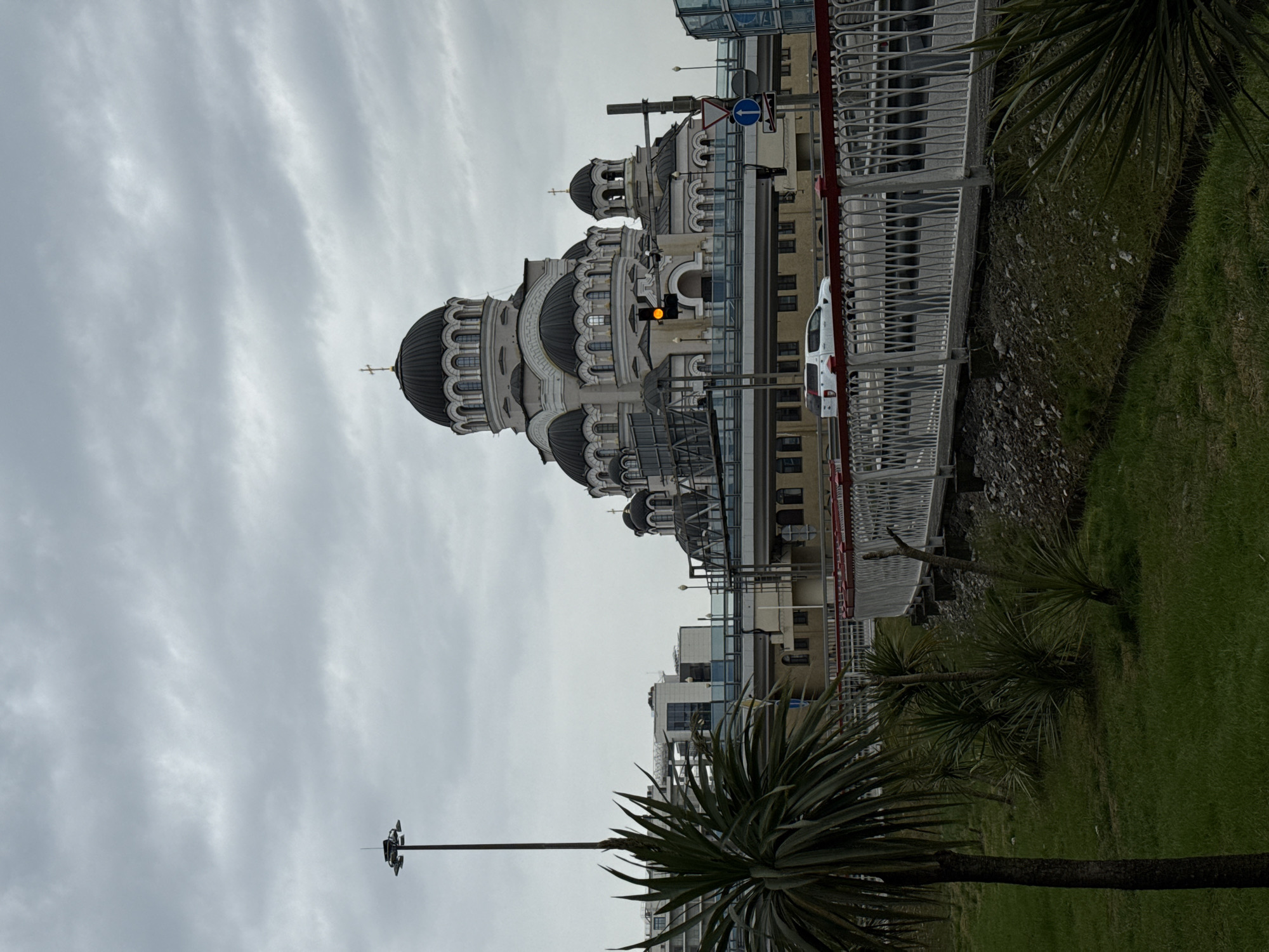 The image depicts a cityscape under a cloudy sky. The most prominent feature is a large, white cathedral with multiple domes topped with crosses. The building's architecture is detailed with rows of windows and decorative elements.
Next to the cathedral, modern buildings with glass and steel structures are visible. A white car is parked on the street. A metal fence runs along a raised platform or walkway.
In the foreground, there's greenery, including several palm-like trees. A lamppost is also visible.
This scene is likely in Sirius, Russia, as stated in the prompt.