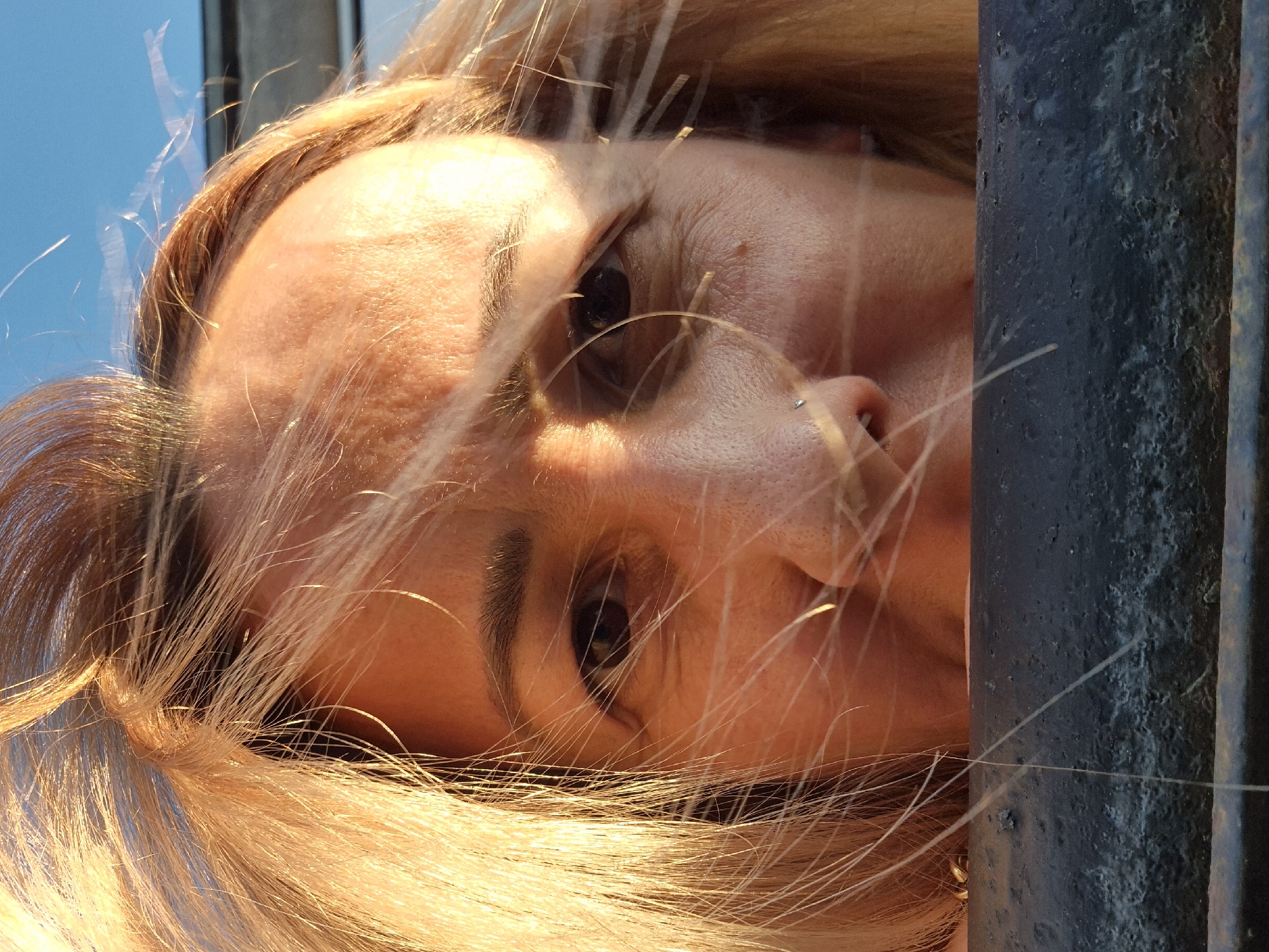 A close-up, low-angle shot captures a woman's face, with her blonde hair blowing across her forehead. The sunlight illuminates the right side of her face, creating a warm glow and casting shadows. Her eyes are dark and looking slightly off-camera. A small nose piercing is visible on her left nostril. Her lips are slightly parted, revealing a glimpse of her teeth. She is positioned behind a dark, textured railing or ledge that occupies the bottom portion of the frame. The background shows a clear, pale blue sky. The scene appears to be outdoors, possibly in a park or elevated area, given the sky and the railing. The image is focused tightly on the woman's face and the foreground railing.