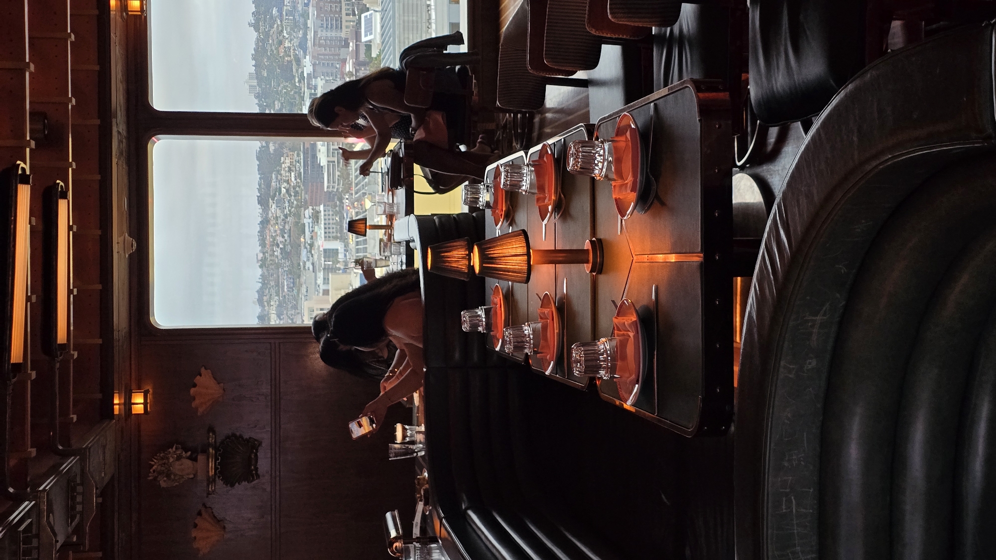 The image depicts the interior of a restaurant with a large window offering a panoramic view of a city skyline. The view through the window suggests the location is San Diego, USA, with buildings and hills visible in the distance under an overcast sky.
Inside the restaurant, a long table is set for dining, with plates, glasses, and small lamps. The table is positioned in front of a dark, curved booth. Several people are seated at tables, some of whom are looking at their phones. The lighting inside is dim and warm, with the table lamps casting an orange glow. The decor appears to be somewhat upscale, with dark wood paneling and ornate wall decorations visible on the left side of the image. Two women are seated at a table by the window, one holding a phone and the other appearing to eat or drink. In the foreground, the dark leather of the booth and chairs is prominent. The overall atmosphere is intimate and sophisticated.