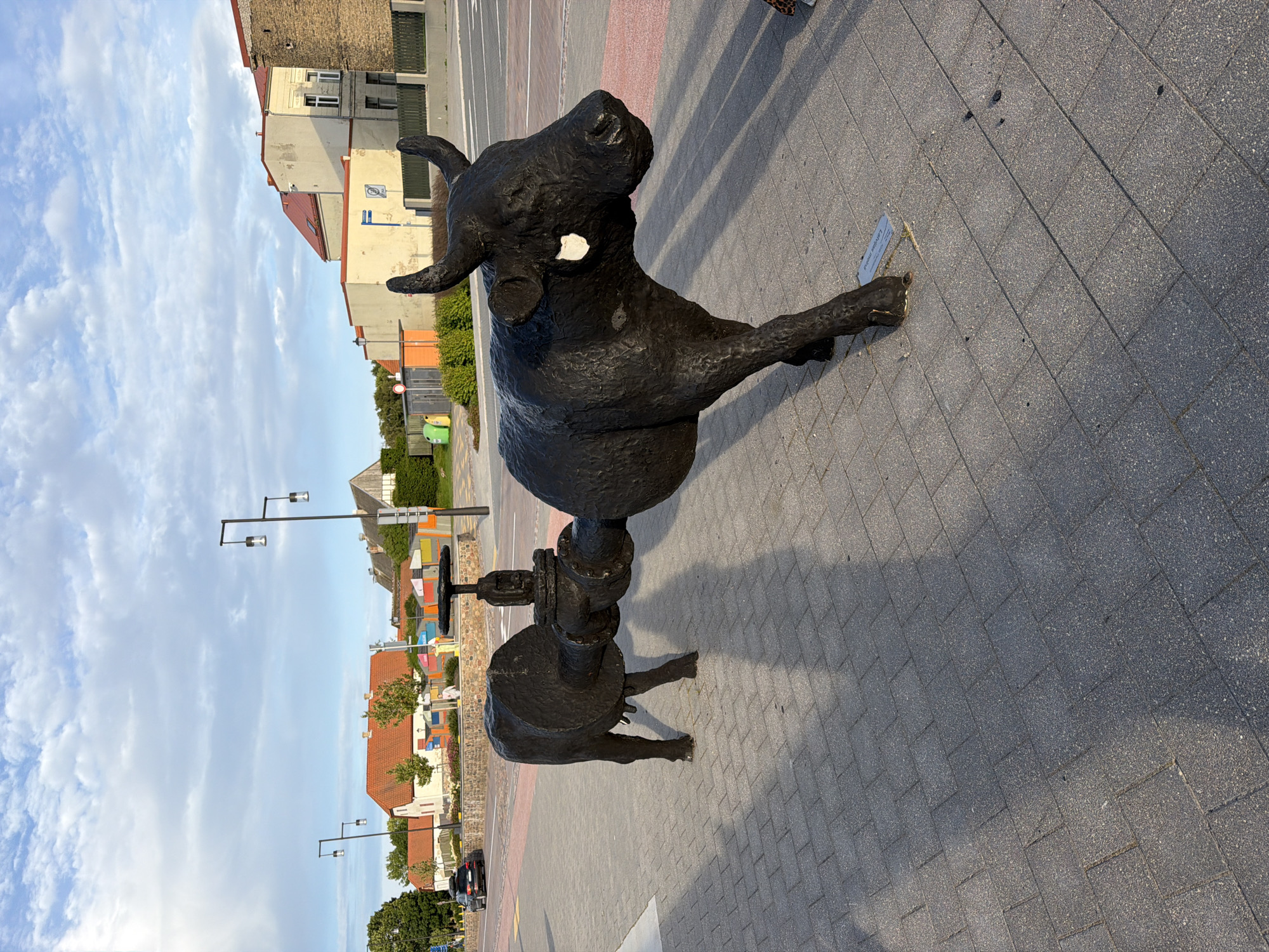 A black sculpture of a bull, with a large pipe passing through its body, stands on a brick-paved sidewalk in Ventspils, Latvia. The bull sculpture is facing to the right, with its head and front legs visible on the right side of the frame, and its hindquarters and back legs on the left. A large valve mechanism is attached to the pipe where it passes through the bull. The background features buildings with red roofs, trees, and a cloudy sky. A car is visible on a street to the left of the sculpture. The lighting suggests it is late afternoon or early evening, with long shadows cast by the sculpture. A small plaque is visible at the base of the bull's front right leg.