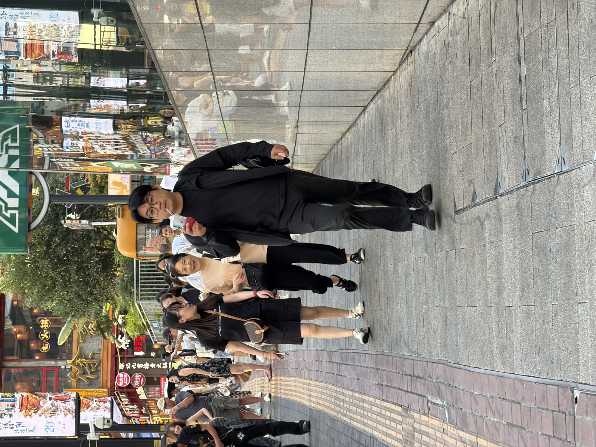 In Yuzhong District, China, a man in a black jacket and pants walks down a pedestrian walkway while drinking a pink beverage from a clear cup with a straw. He is wearing glasses and has dark hair. To his left, two women walk in the same direction; one in a black t-shirt and shorts, carrying a small brown handbag, and the other in a tan t-shirt and dark pants, carrying a black bag. Other pedestrians, buildings with illuminated signs, and a yellow vehicle are visible in the background. The walkway is paved with grey stone tiles, and there is a textured yellow and red tiled curb to the left.