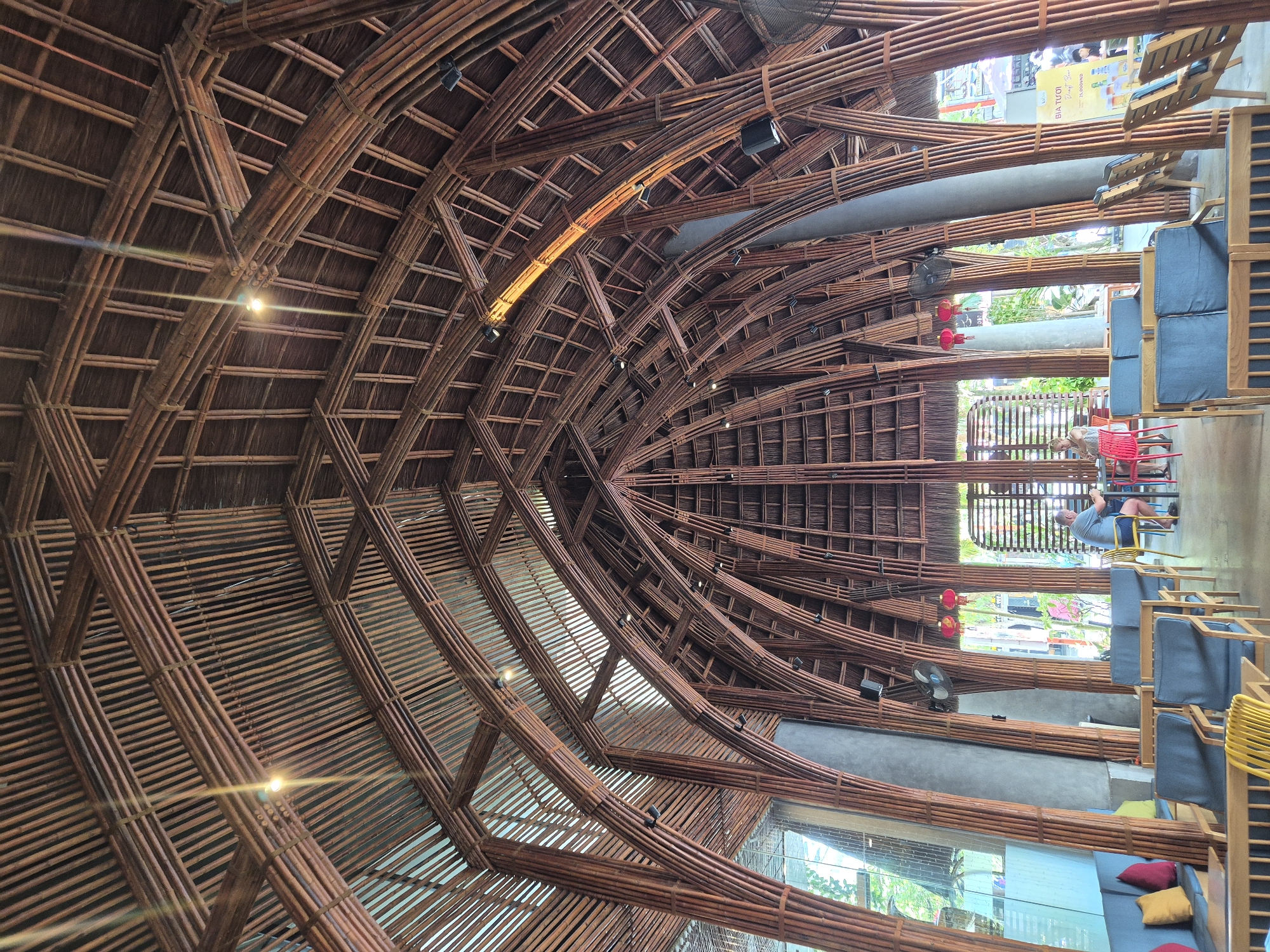 The image depicts the interior of a building with a distinctive bamboo structural design, likely a cafe or restaurant in Đà Nẵng, Vietnam. The ceiling is an elaborate construction of interlaced bamboo poles forming a vaulted roof, with visible lighting fixtures and what appears to be thatched roofing material. The main structure is supported by numerous thick, vertical bamboo columns that arch upwards and outwards.
In the foreground, there are seating areas with wooden chairs and blue upholstered sofas, some with colorful accent pillows. The environment suggests a public space designed for relaxation and socializing.
In the mid-ground, a man and a woman are seated at a table, engaged in conversation or a meal. The man is wearing a blue t-shirt and shorts, and the woman is wearing a white dress. They are the primary human subjects in the scene.
The background reveals more of the bamboo architecture and glimpses of the outside environment through large windows or open spaces, suggesting a tropical or semi-tropical setting with vegetation visible. Decorative red lanterns are suspended from the ceiling. A promotional poster is visible on a pillar in the right side of the image. The overall impression is one of an architecturally unique and inviting indoor space.